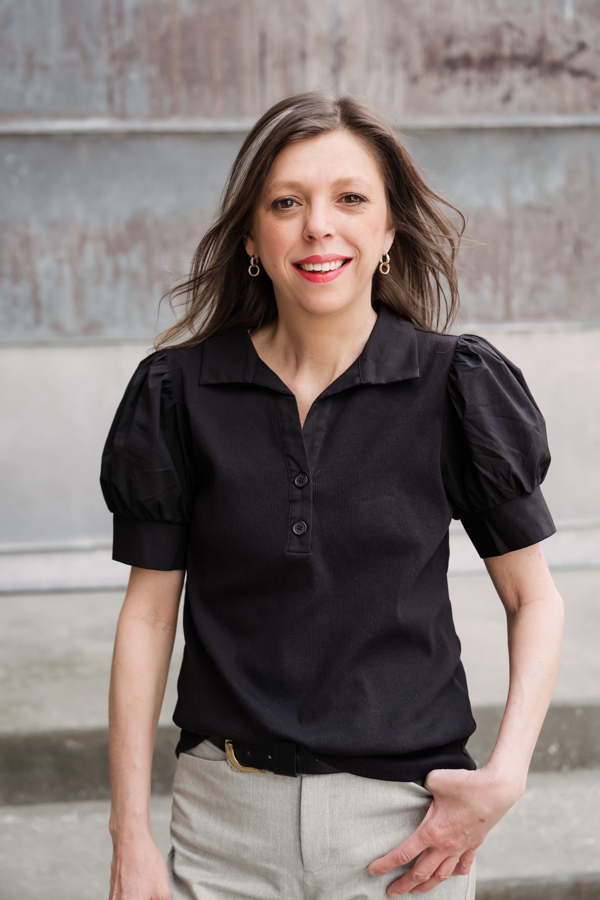 Woman standing outdoors in a black blouse and light pants, smiling against a concrete wall.