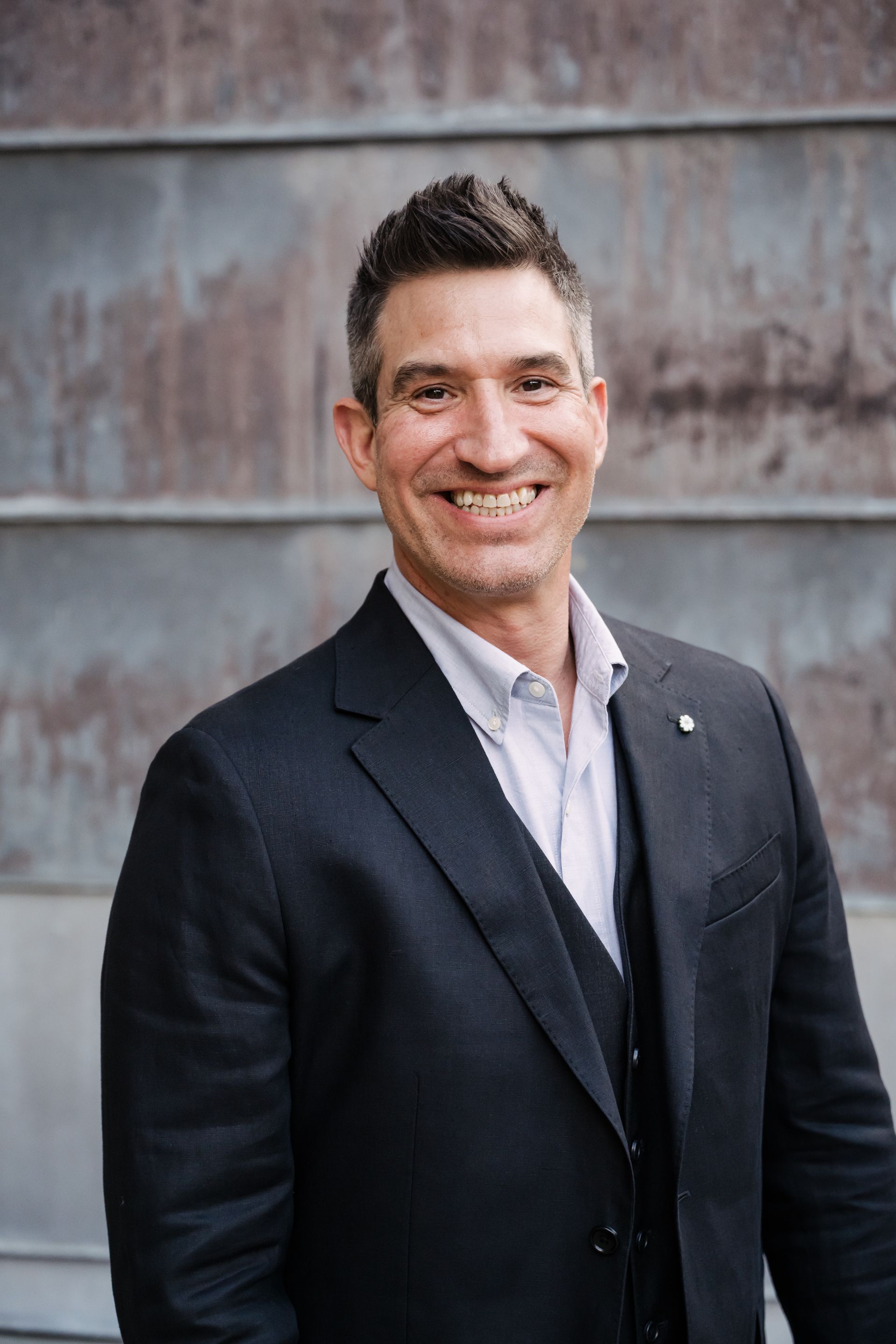 Smiling man in a dark suit standing against a weathered metal wall