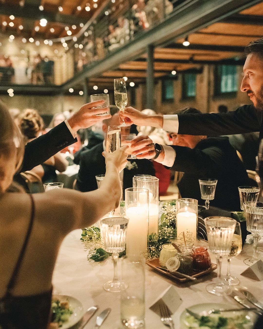 Guests toast with glasses at a warmly lit dinner table in a festive indoor venue.