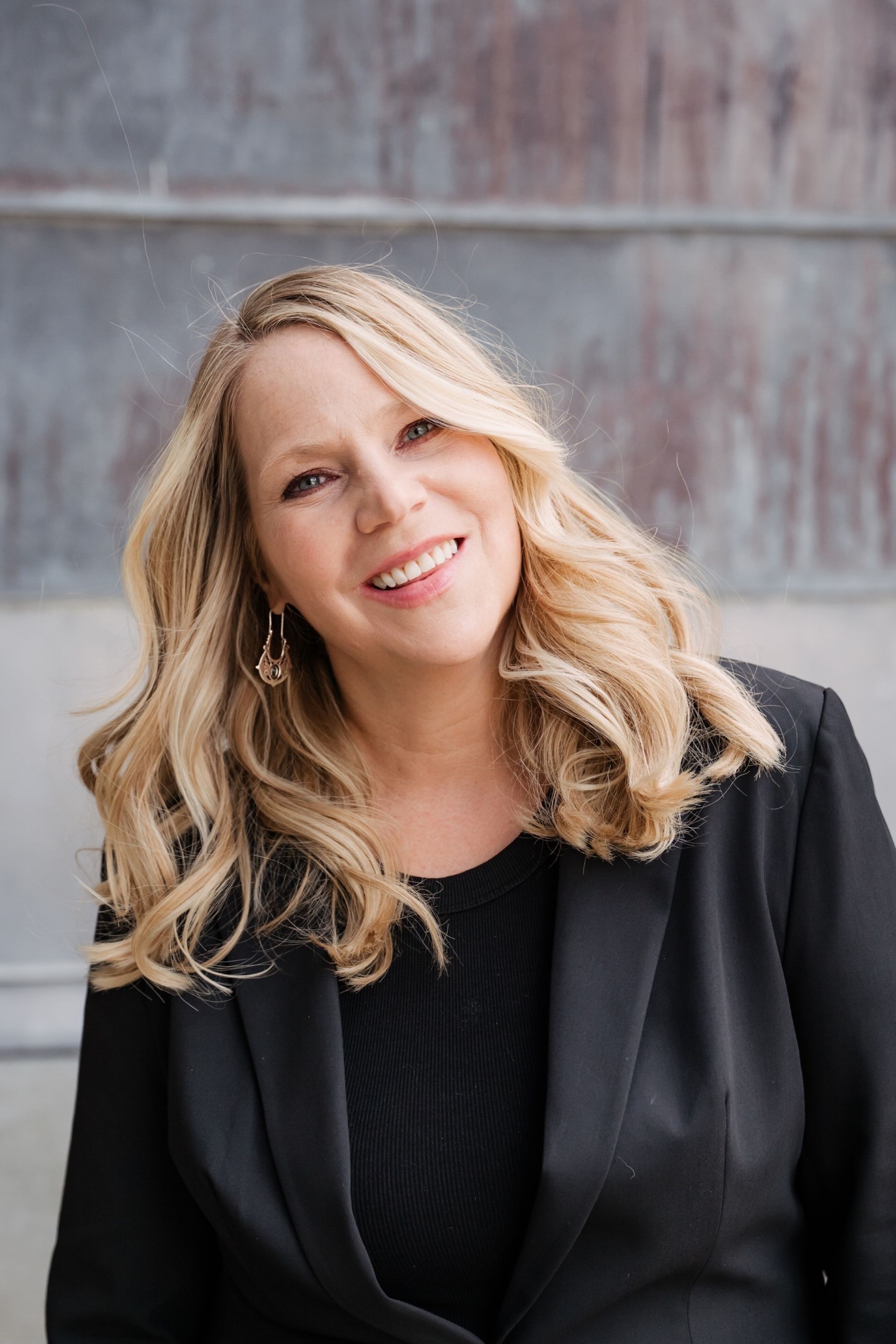 Smiling woman with blonde hair in a black jacket against a gray background
