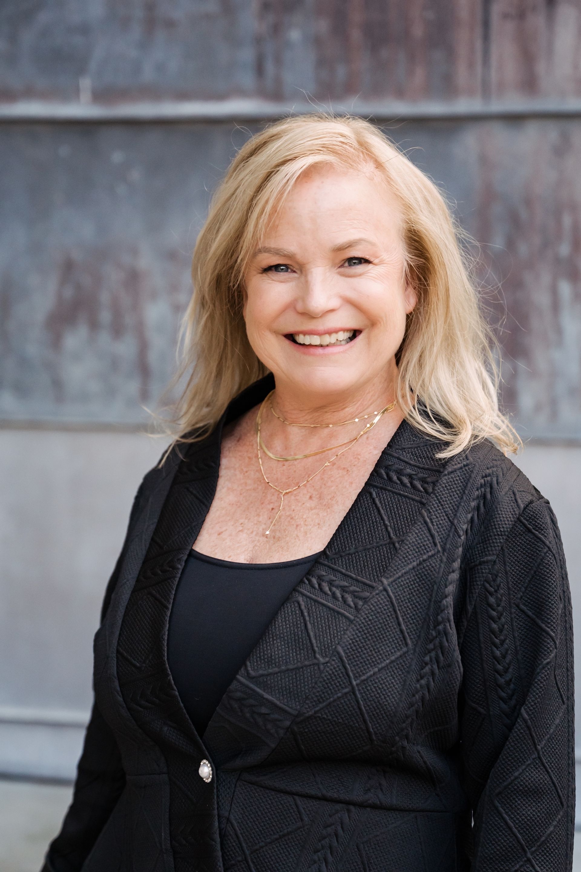 Smiling woman in a black jacket standing against a gray wall