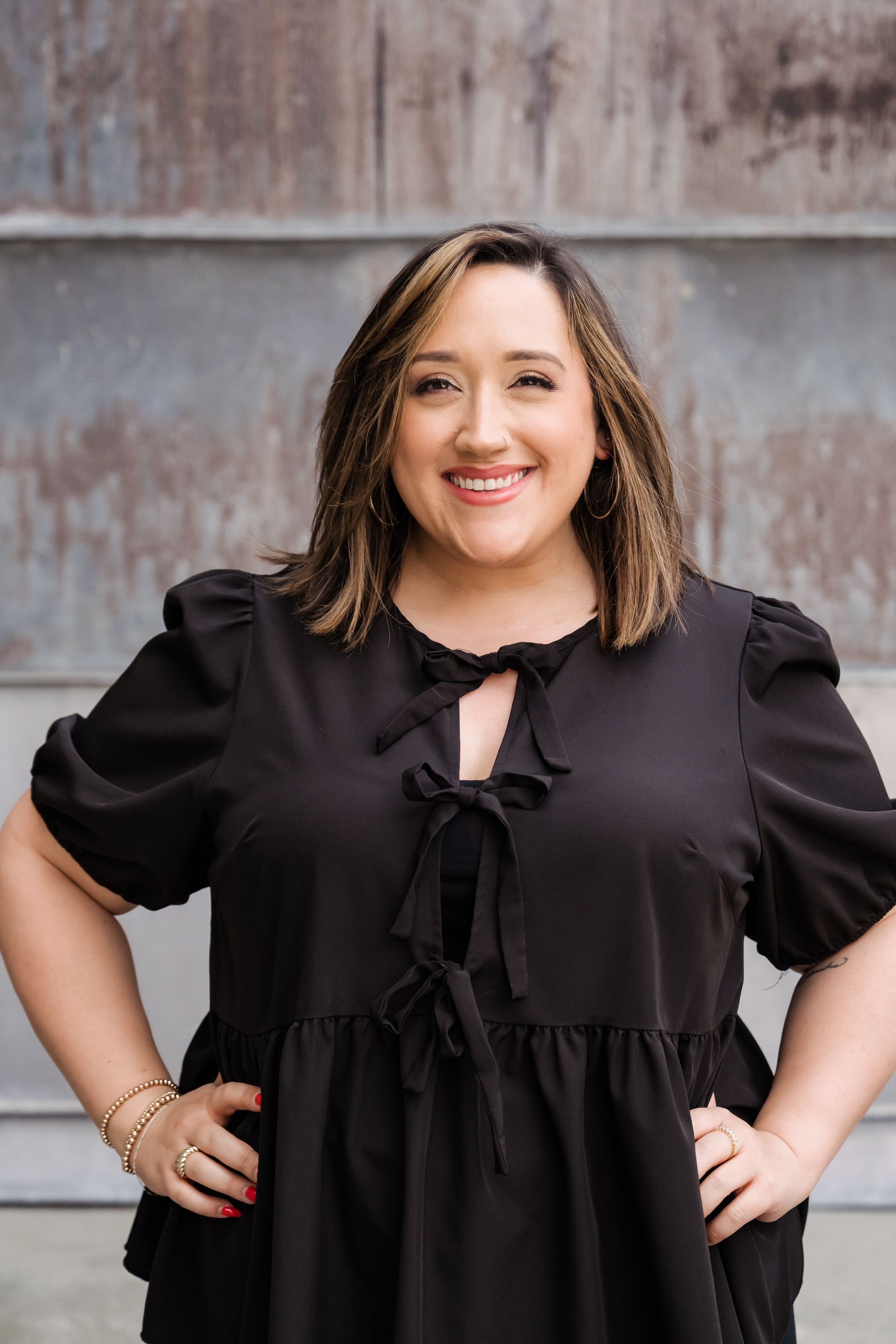 Smiling person in a black dress with hands on hips, standing before a textured gray wall.