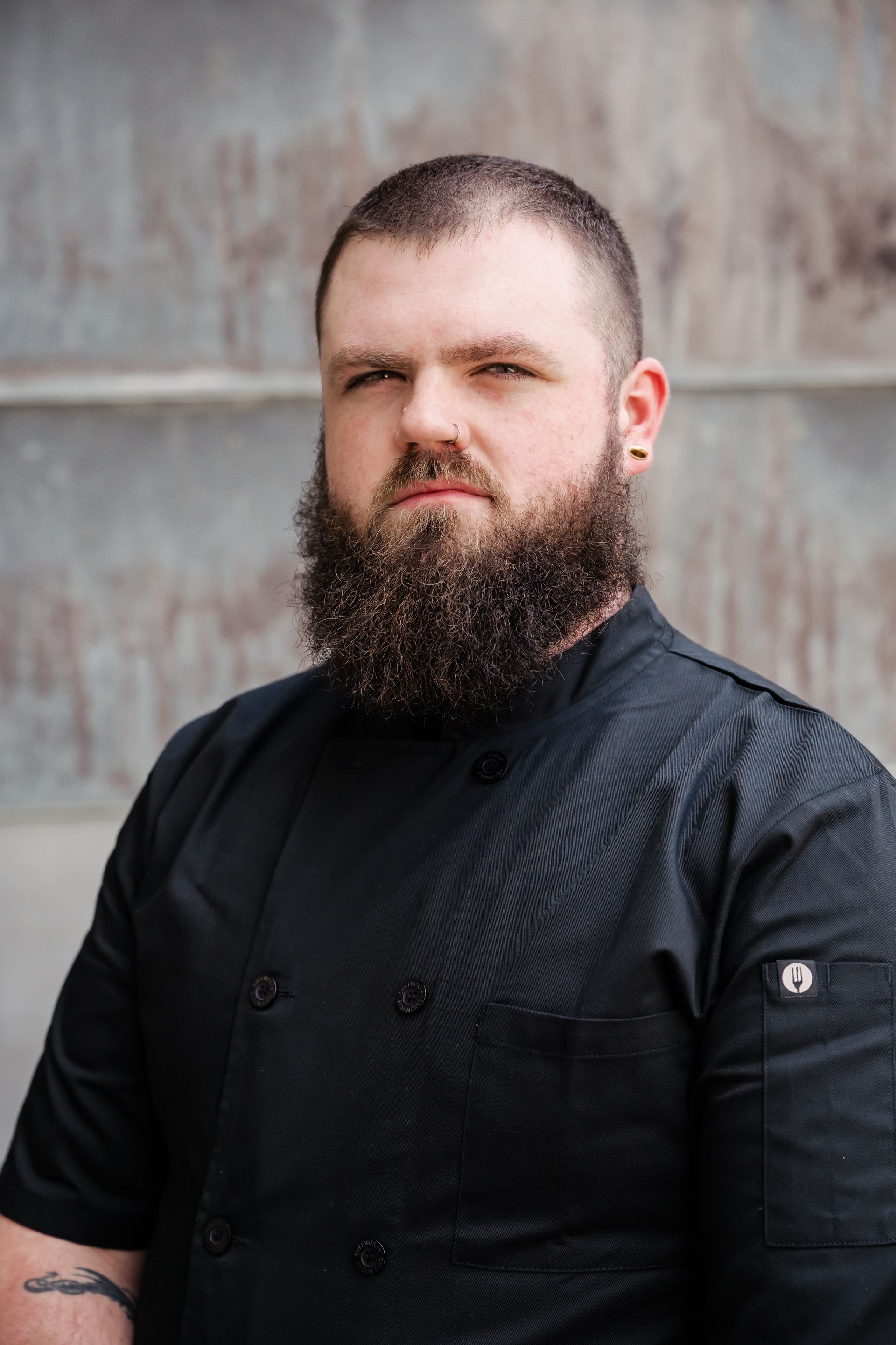 Bearded man in a black shirt posing against a weathered wall