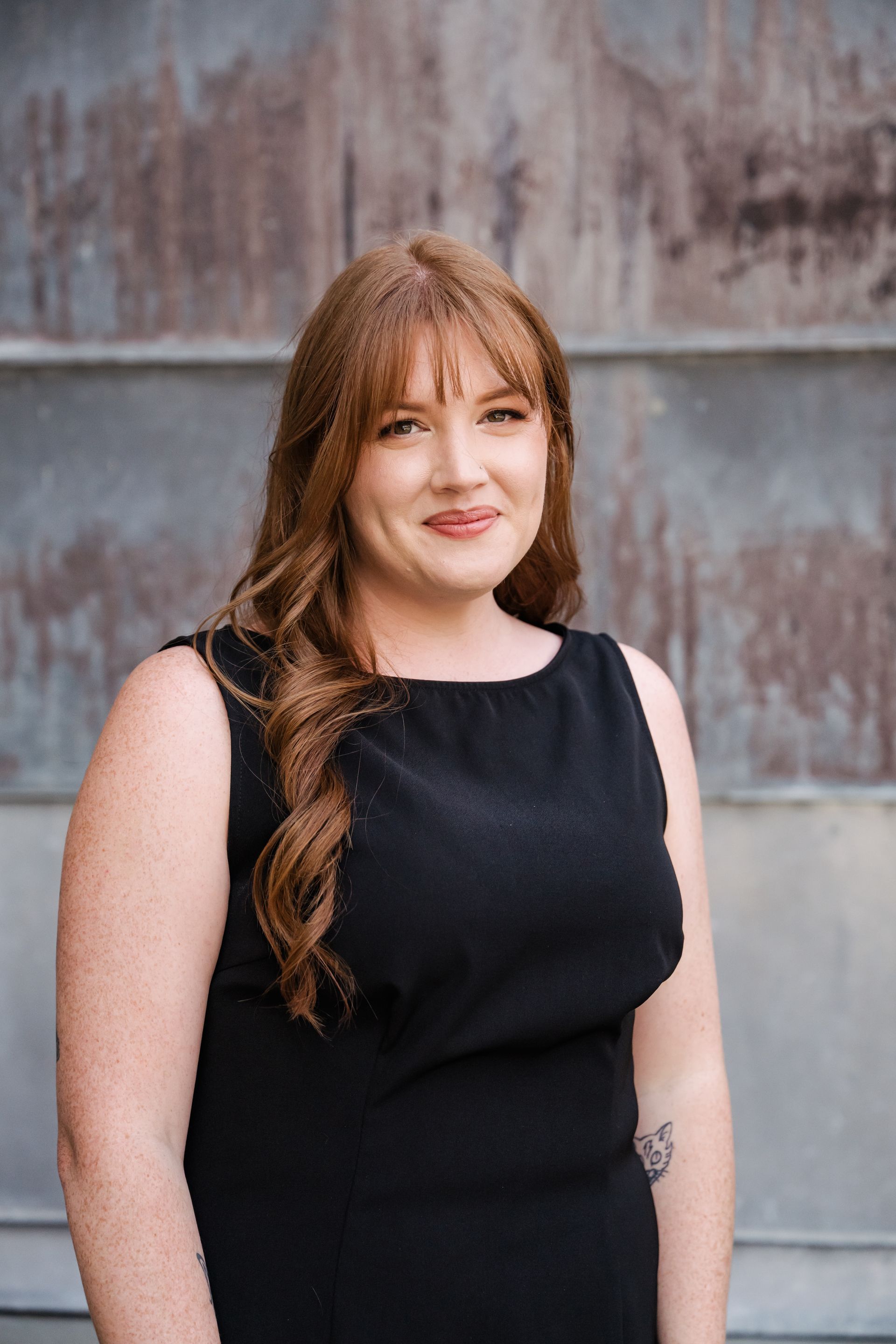 Woman in a black sleeveless dress standing against a weathered metal wall, smiling at the camera