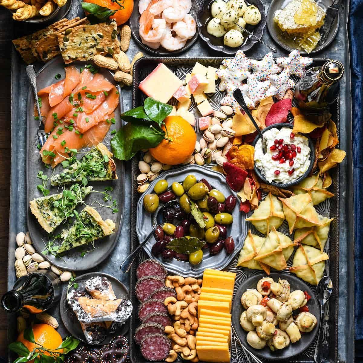 Assorted Mediterranean appetizers and snacks arranged on a dark tray, with olives, cheese, fish, fruit, and crackers
