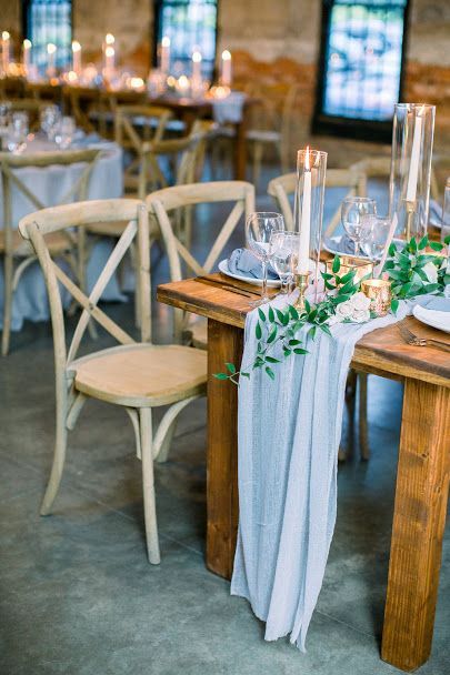 Rustic wedding reception table with beige chairs, greenery, glassware, and a draped gray table runner