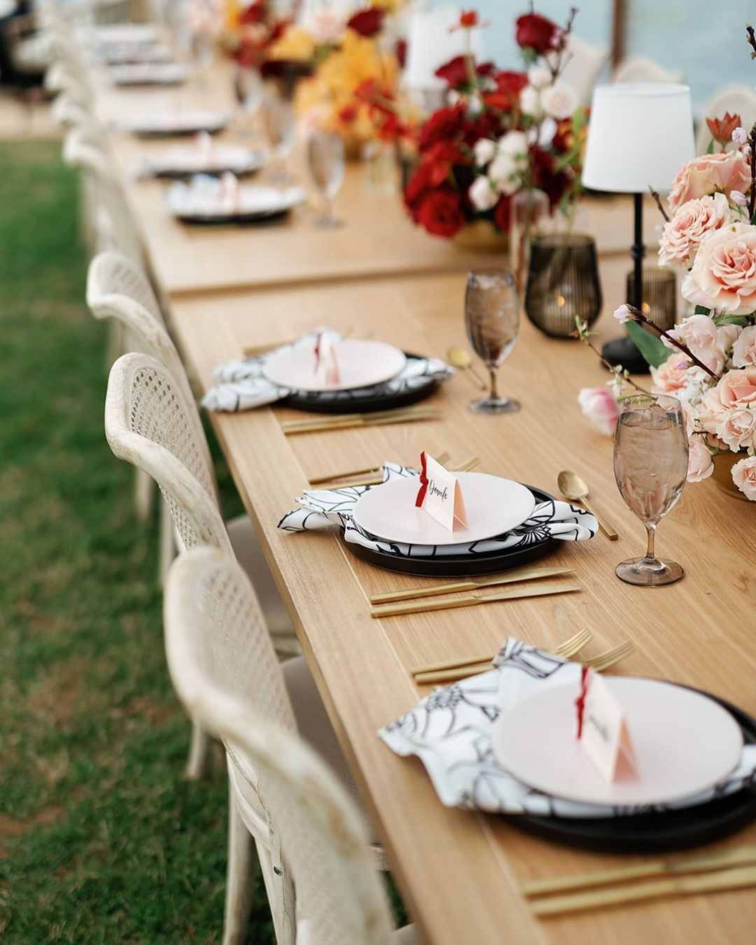 Long outdoor dining table set with white chairs, floral centerpieces, plates, and wine glasses on a lawn