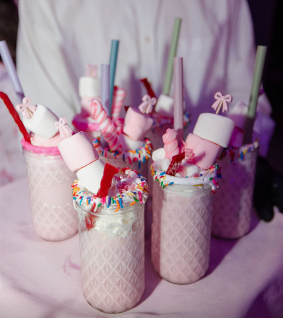 Pink milkshakes topped with whipped cream, marshmallows, and colorful sprinkles in patterned glasses