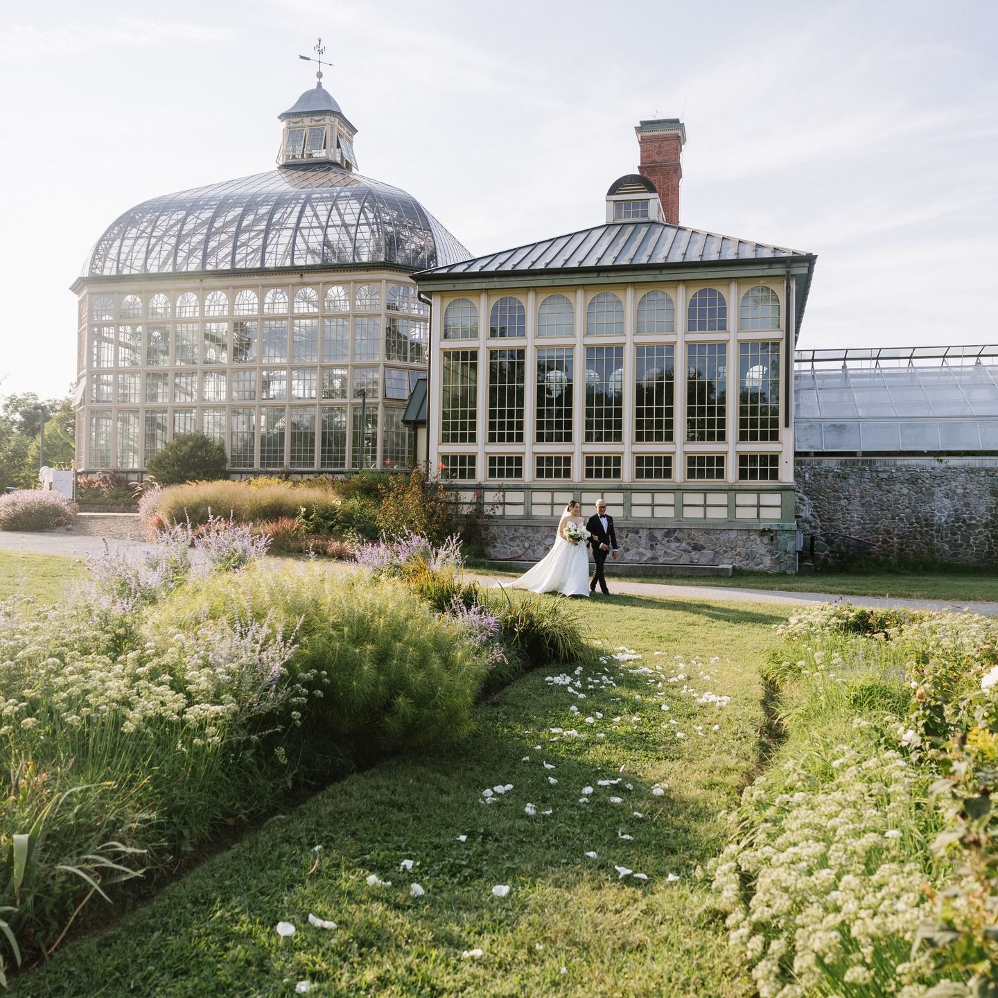 Couple in wedding attire beside a glass conservatory and garden path under soft sunlight