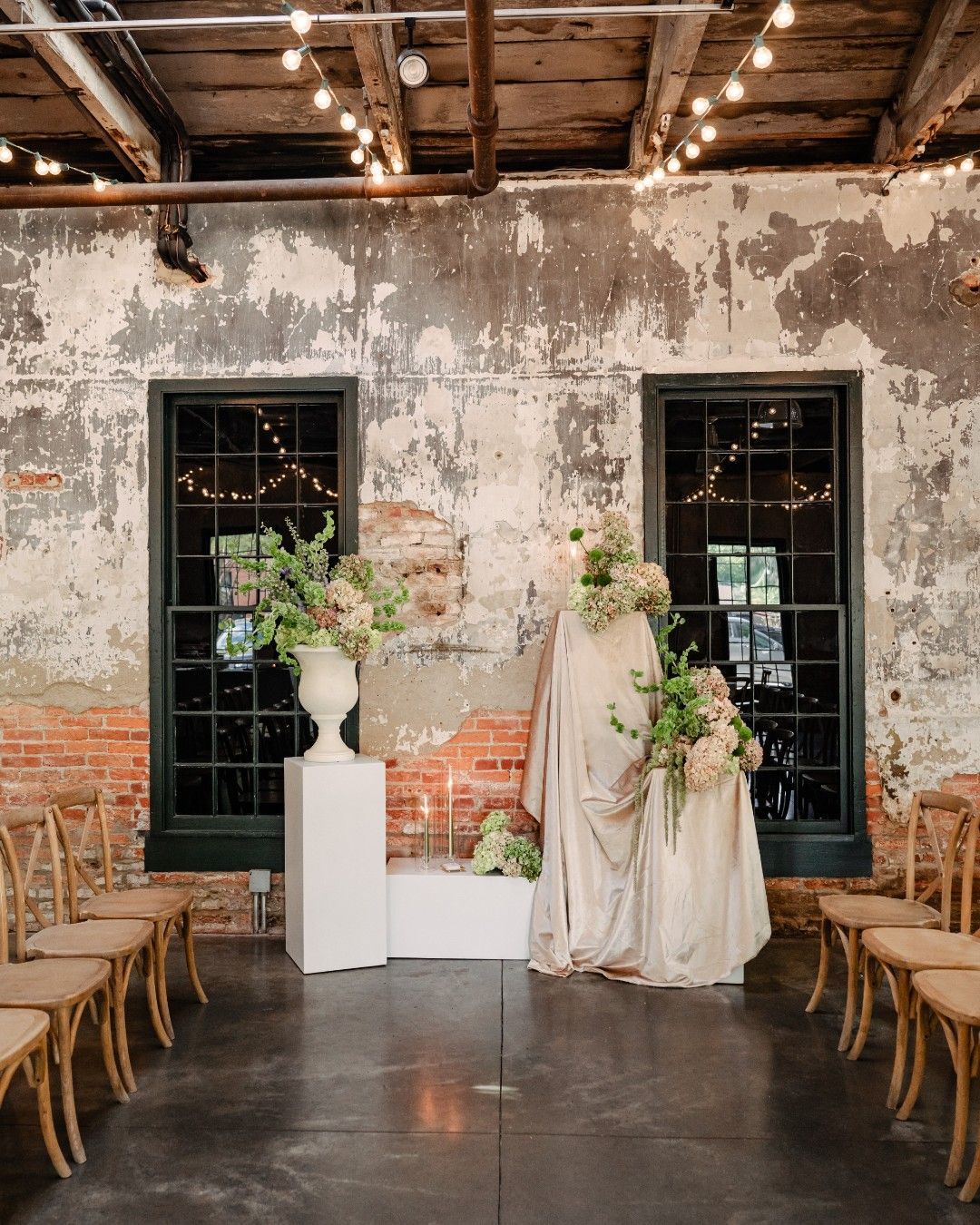 Rustic wedding ceremony setup with floral decor, draped table, and chairs in an industrial hall