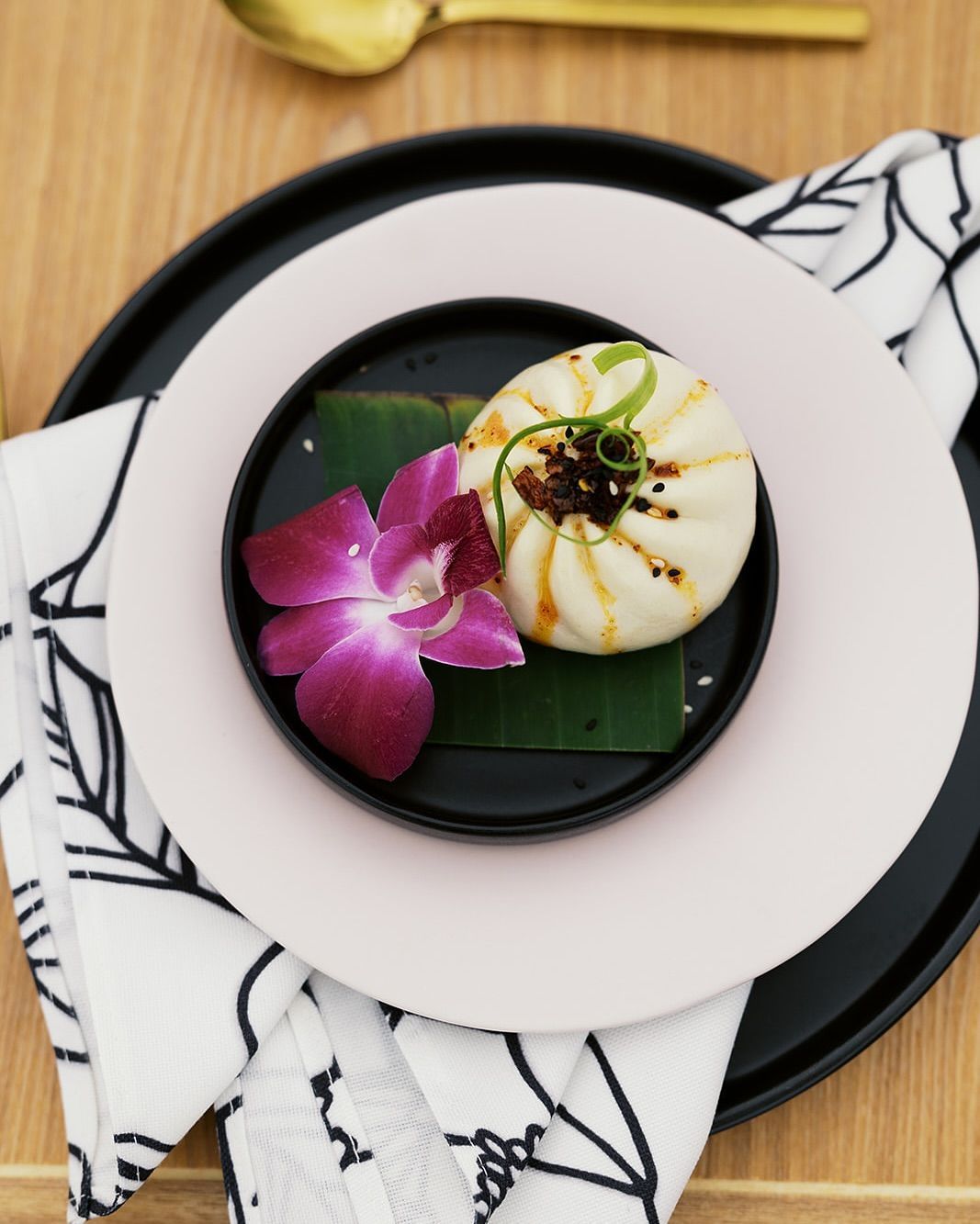 Plated gourmet dish with purple flower garnish and citrus on a white plate over a black plate, on a patterned napkin