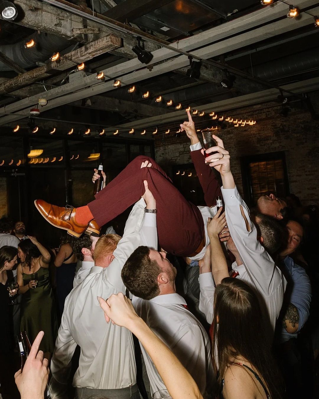 Crowd lifting a person in a white shirt and red pants at a lively indoor party under string lights