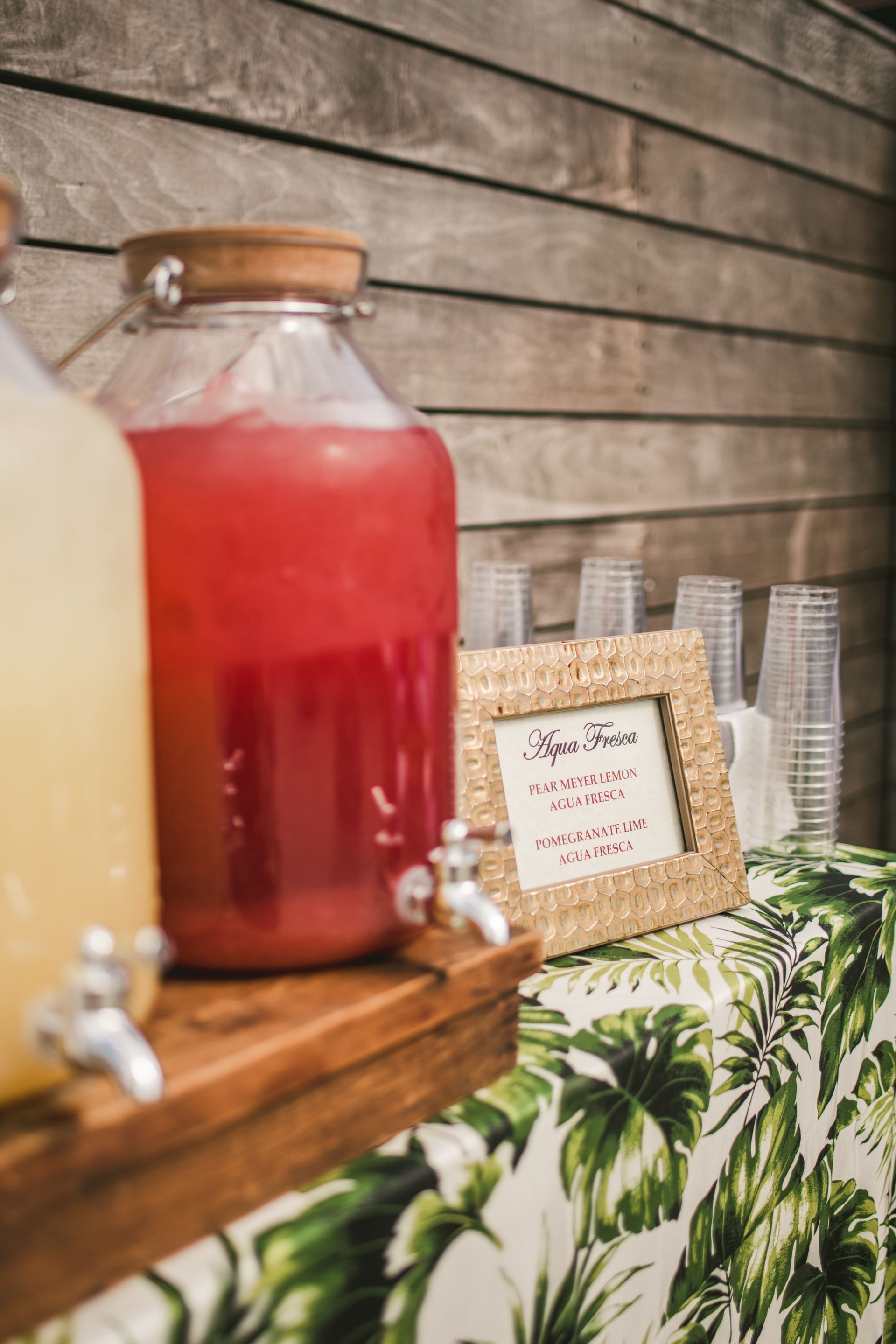 Red beverage in a glass jar beside a framed label and stacked cups on a rustic table