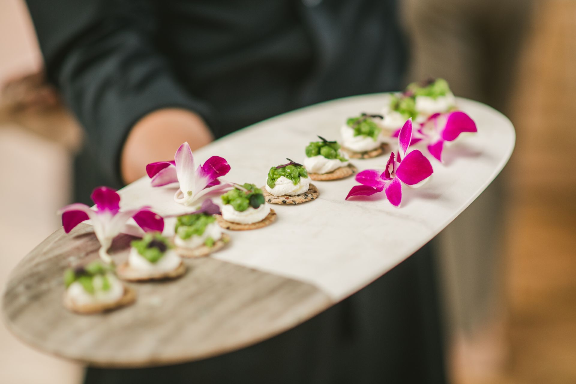Tray of bite-sized hors d'oeuvres garnished with green herbs and purple flowers, held by a person in dark clothing