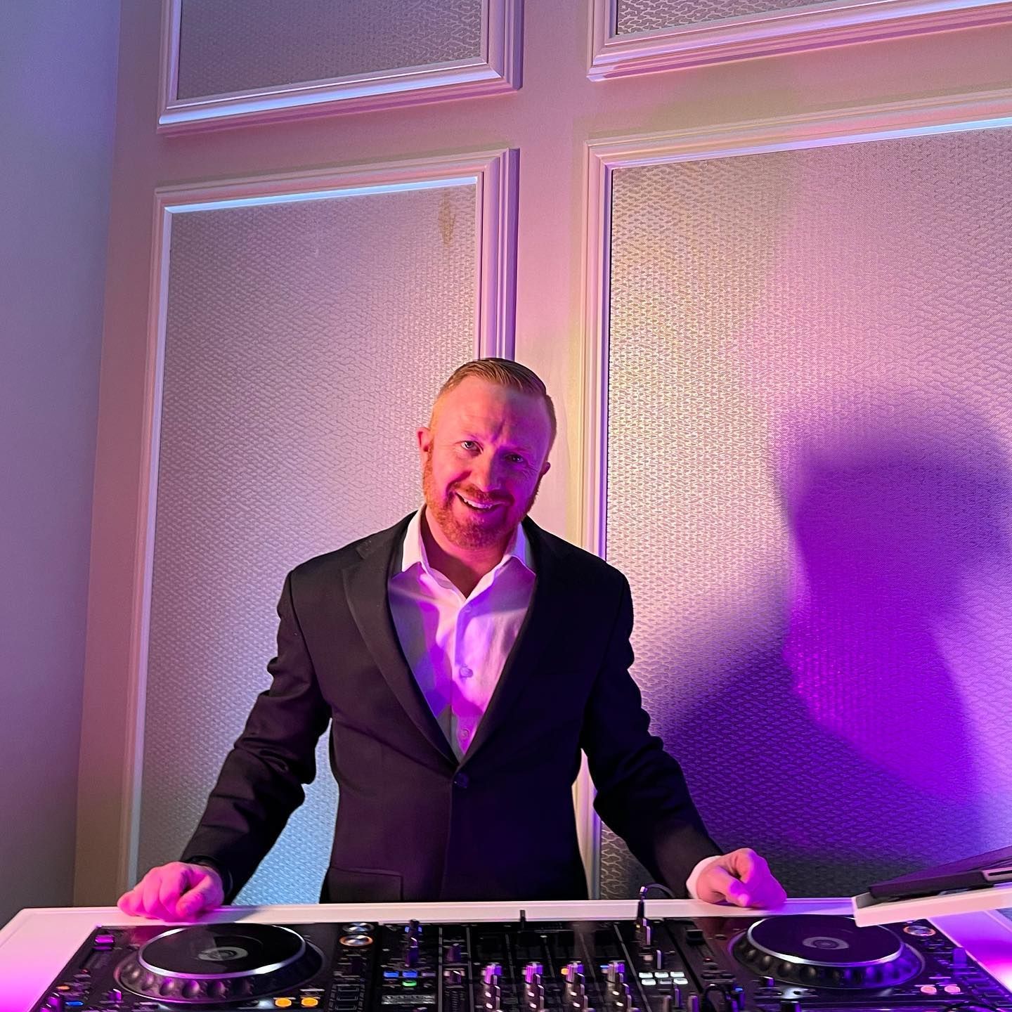 DJ standing behind a mixer in a purple-lit room, hands on the turntables