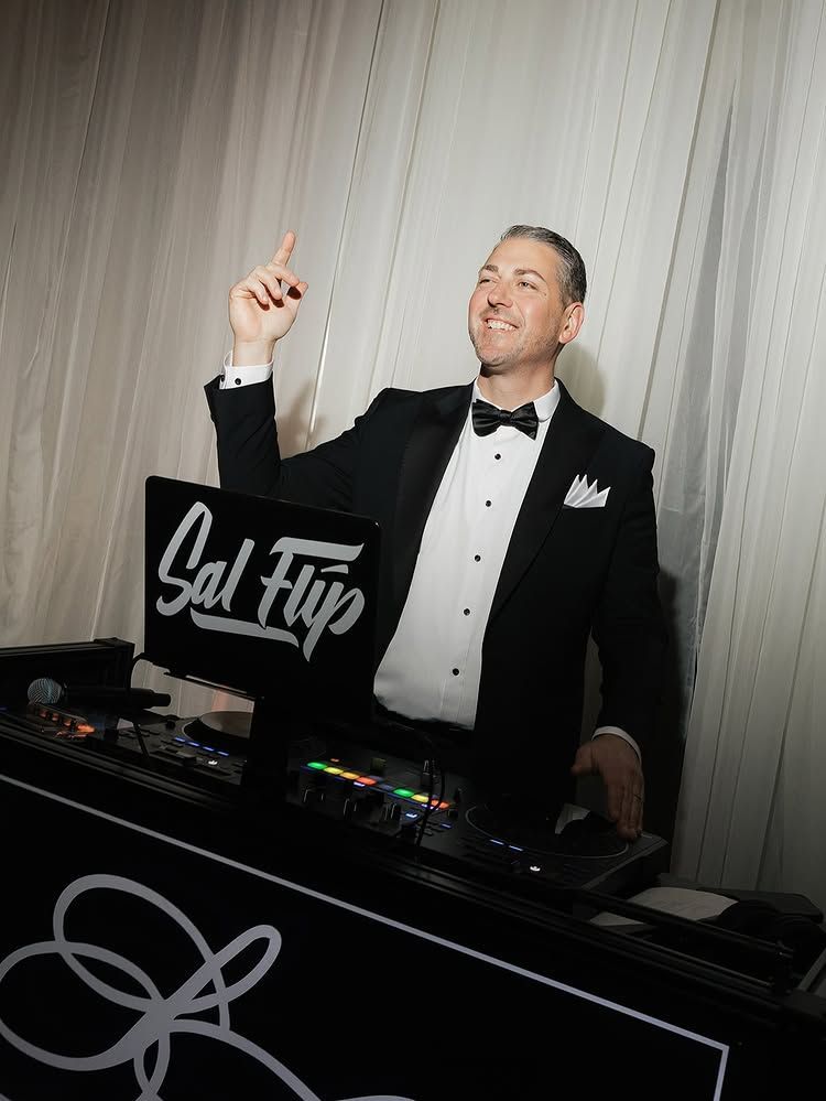 DJ in tuxedo behind a booth, smiling and pointing upward at a black-and-white event setup.