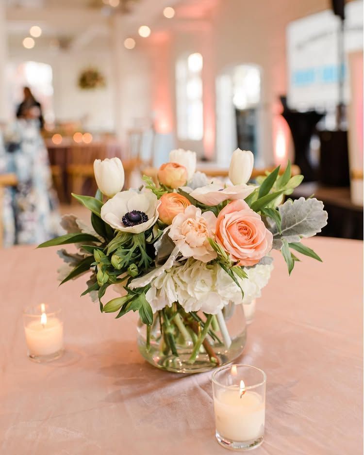 Soft peach and white floral centerpiece in a glass vase on a decorated table with lit candles
