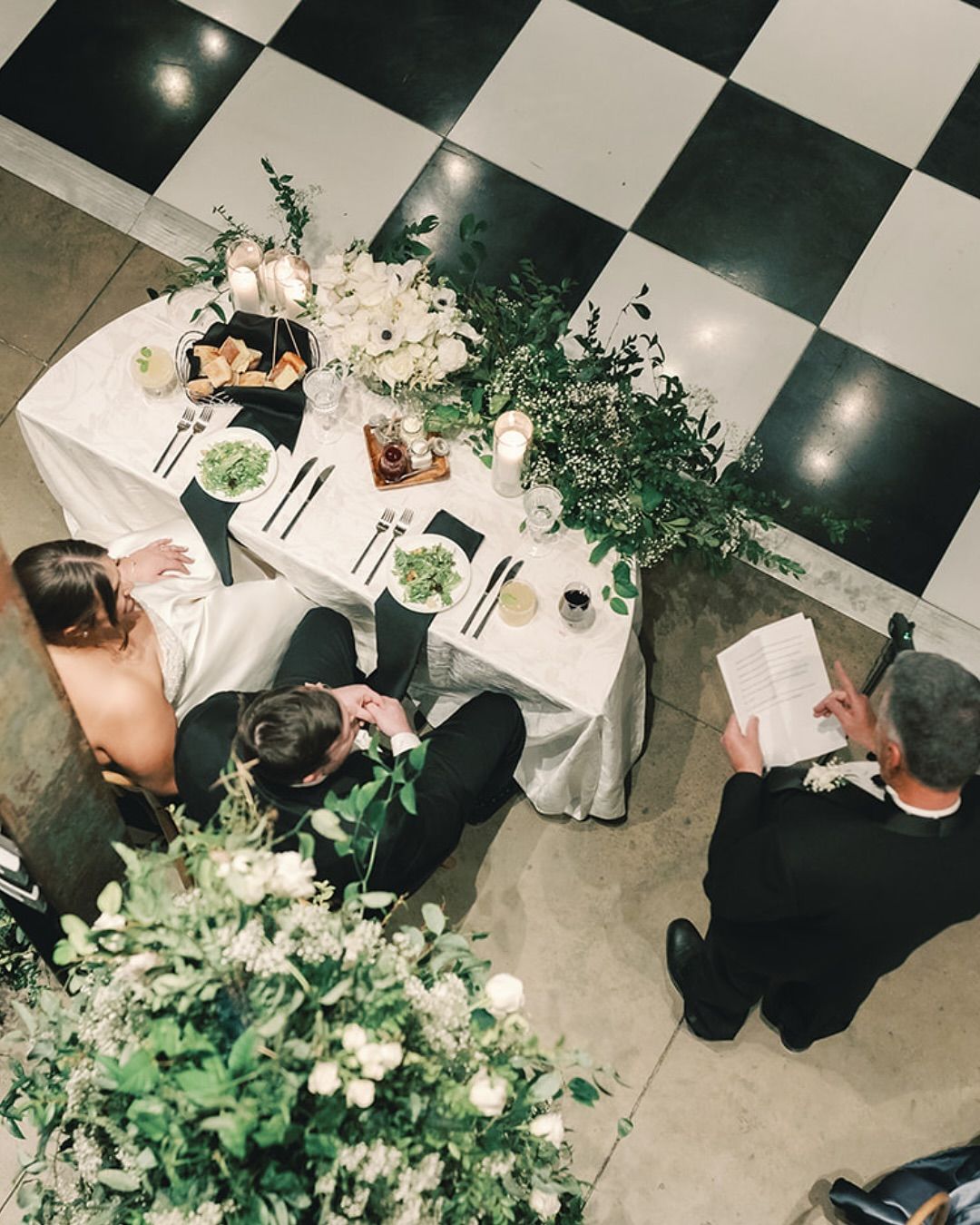 Overhead view of a formal dinner table with flowers, plates, and guests seated on a black-and-white checkered floor