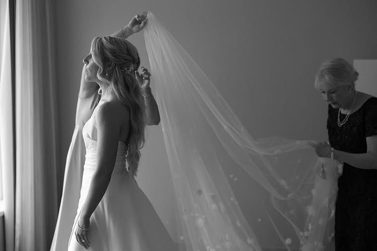 Bride adjusting veil while an assistant arranges the sheer fabric in a softly lit room