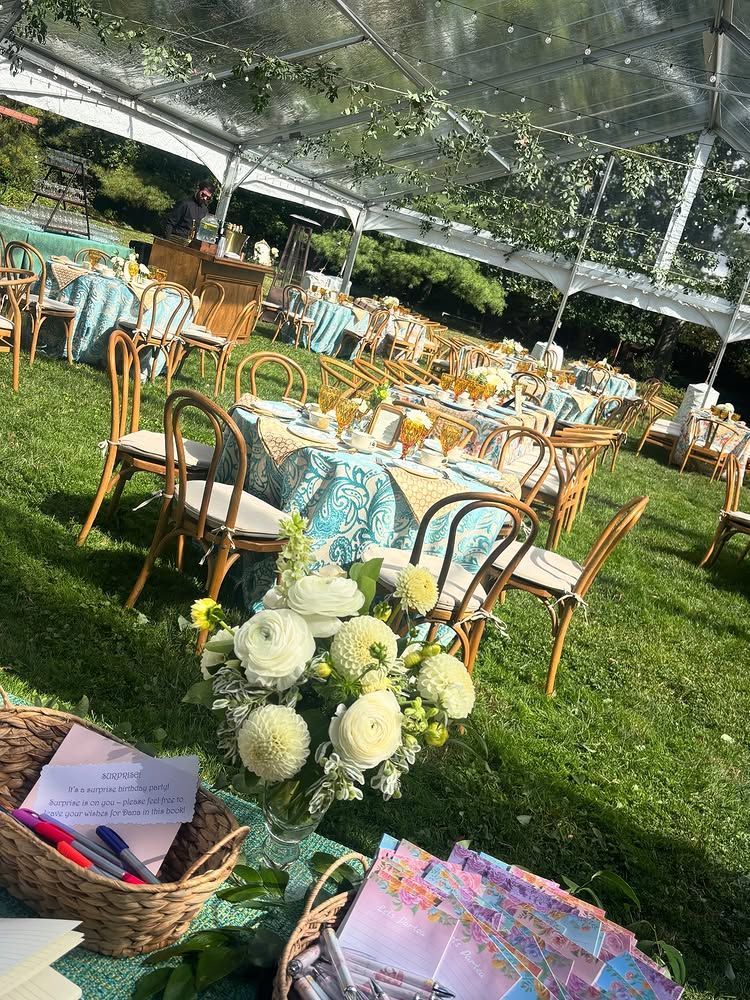 Outdoor wedding reception setup under a clear tent with gold chairs, round tables, and white floral centerpieces on grass