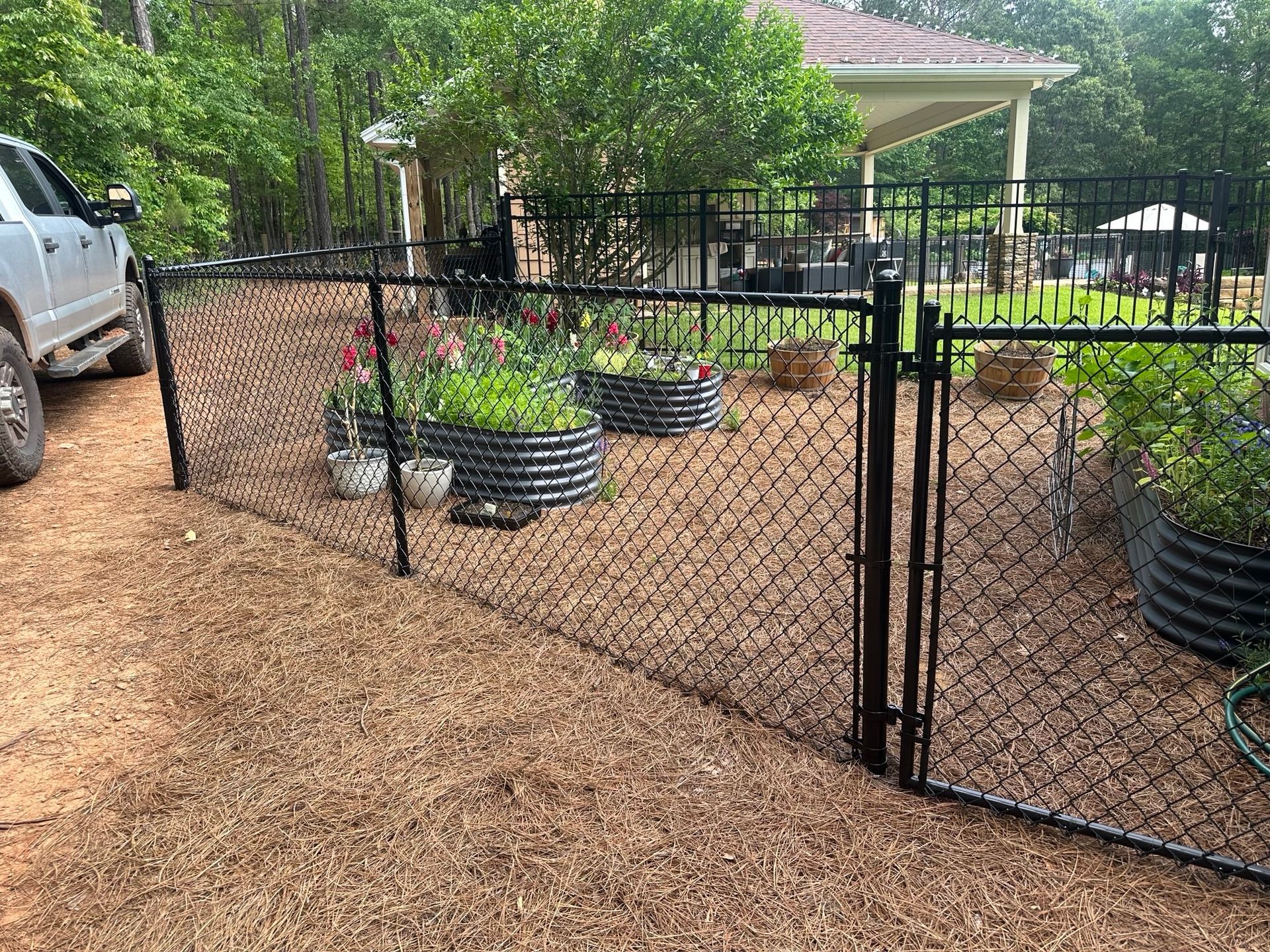 Black chain-link fence encloses a garden with raised beds and flowers; a truck is parked nearby.