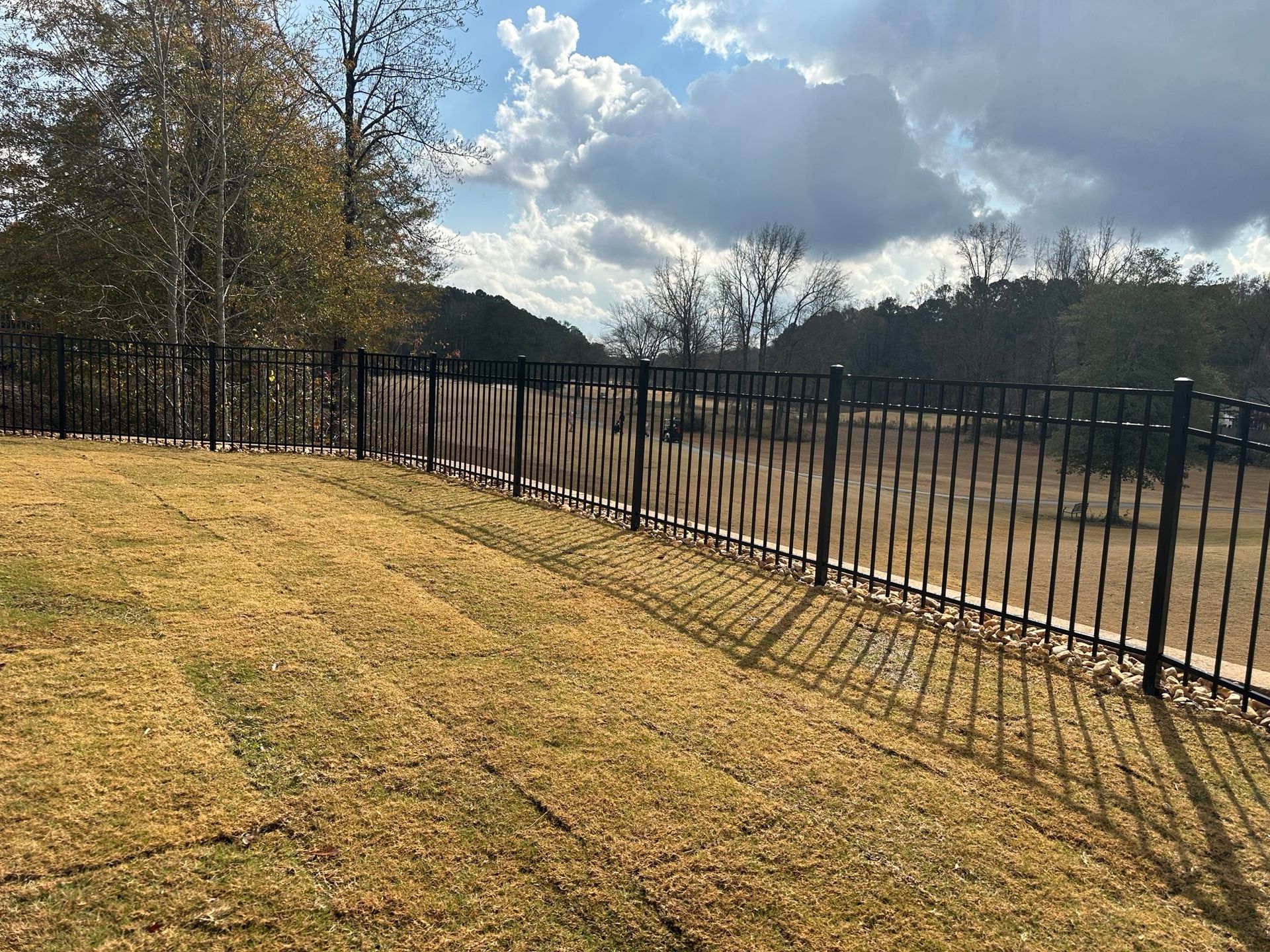 Black metal fence bordering a brown grassy yard under a partly cloudy sky.