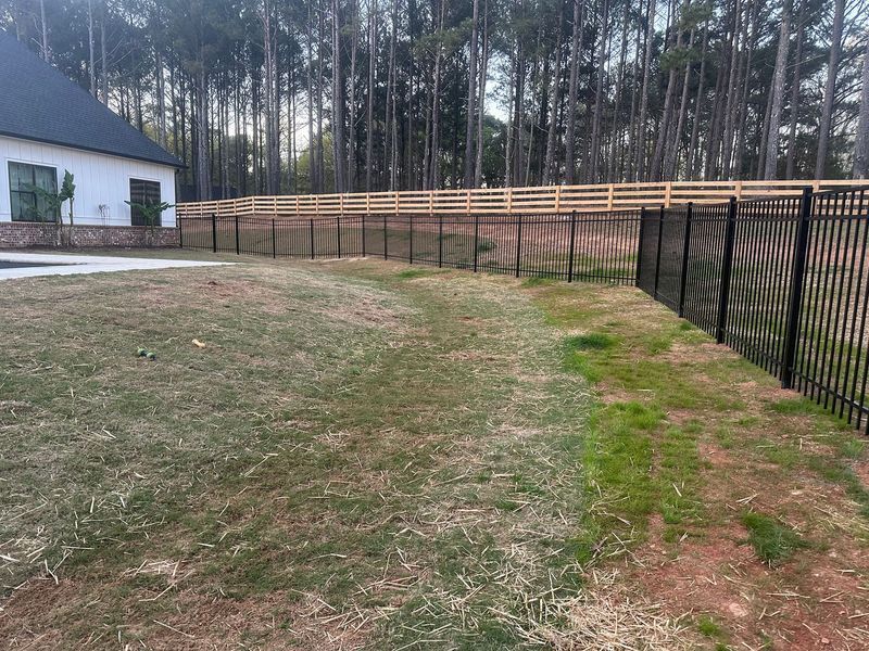 A black metal fence and wooden fence enclose a grassy yard near a white house and a line of trees.