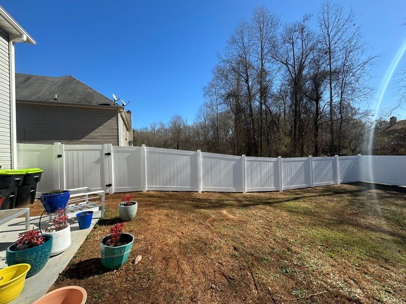 White vinyl fence in a backyard with potted plants and trees in the background. Sunny day.