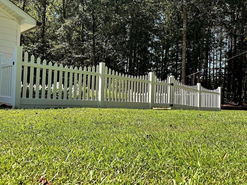 White picket fence in a grassy yard, with a building on the left and trees in the background.