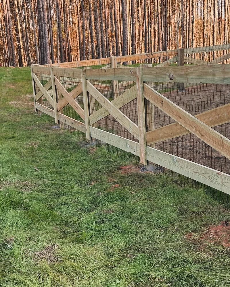 Wooden fence with crisscross design on a grassy hill, forest backdrop.