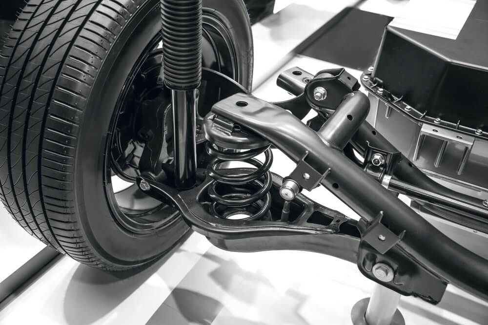 Black and white close-up of a car's rear suspension system with a tire, spring, and control arms visible.