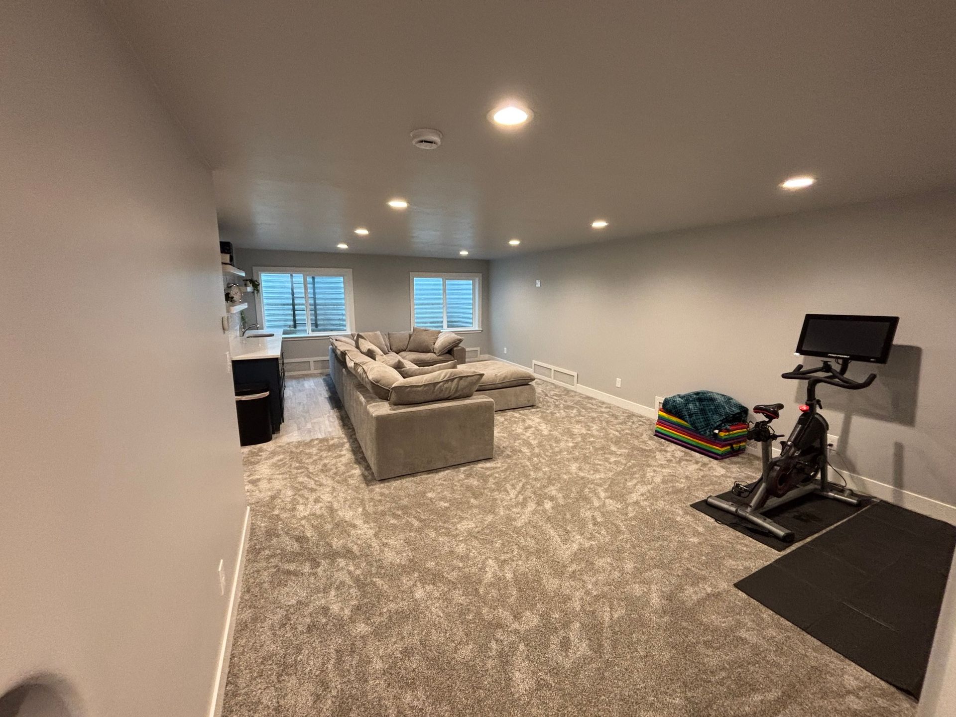 Spacious basement media room with gray sectional sofa, carpet, recessed lights, and exercise equipment by the wall