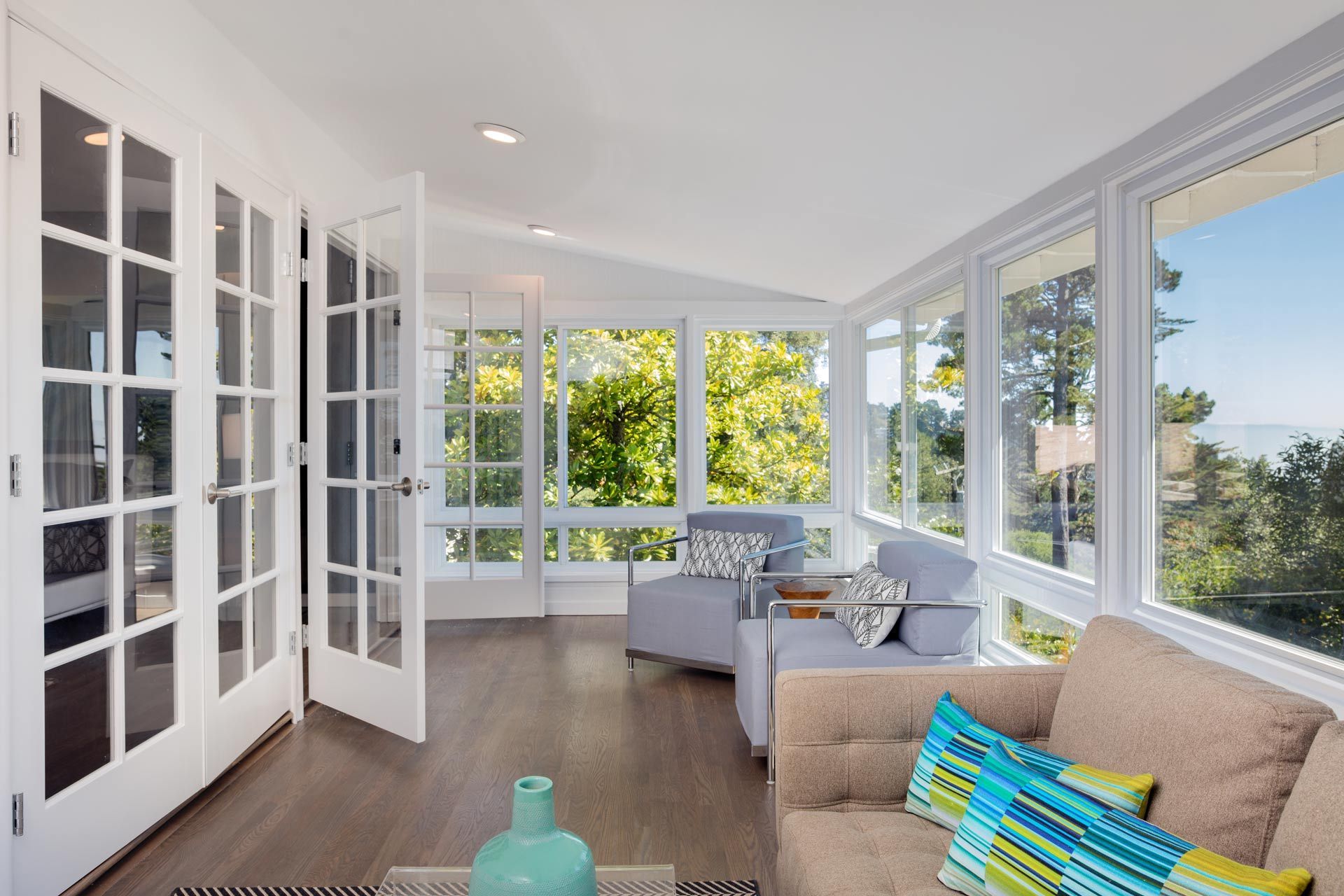 Bright sunroom with white French doors, large windows, beige sofa, and colorful pillows.