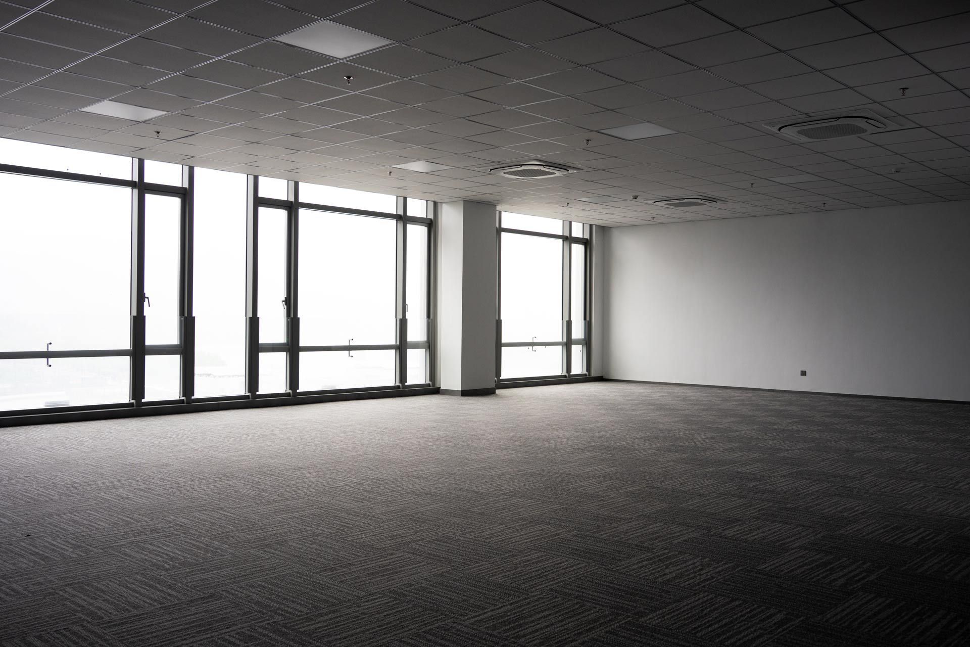 Empty carpeted office space with large windows and overhead fluorescent lighting