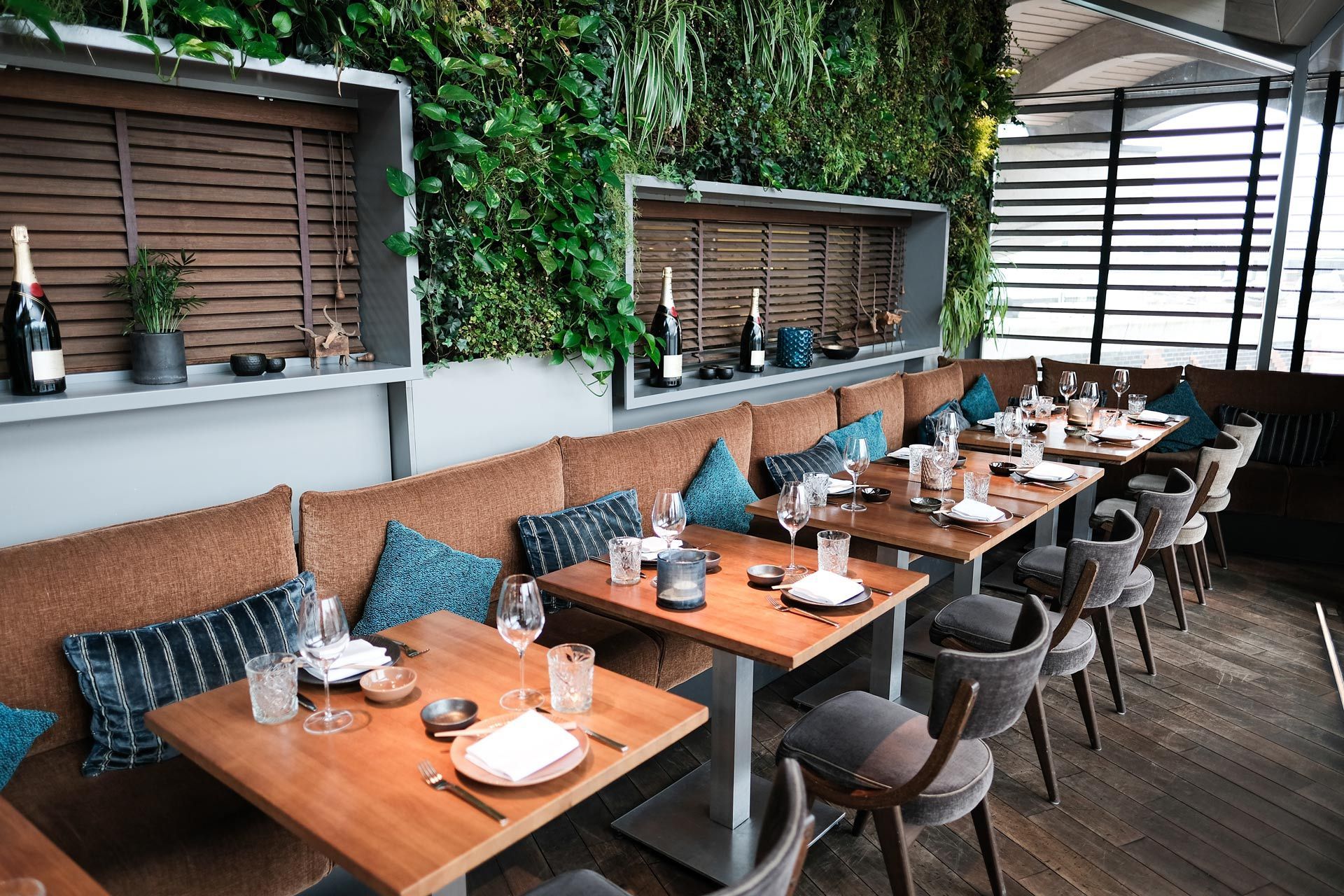 Outdoor restaurant patio with wooden tables, woven chairs, and bottles along a green wall
