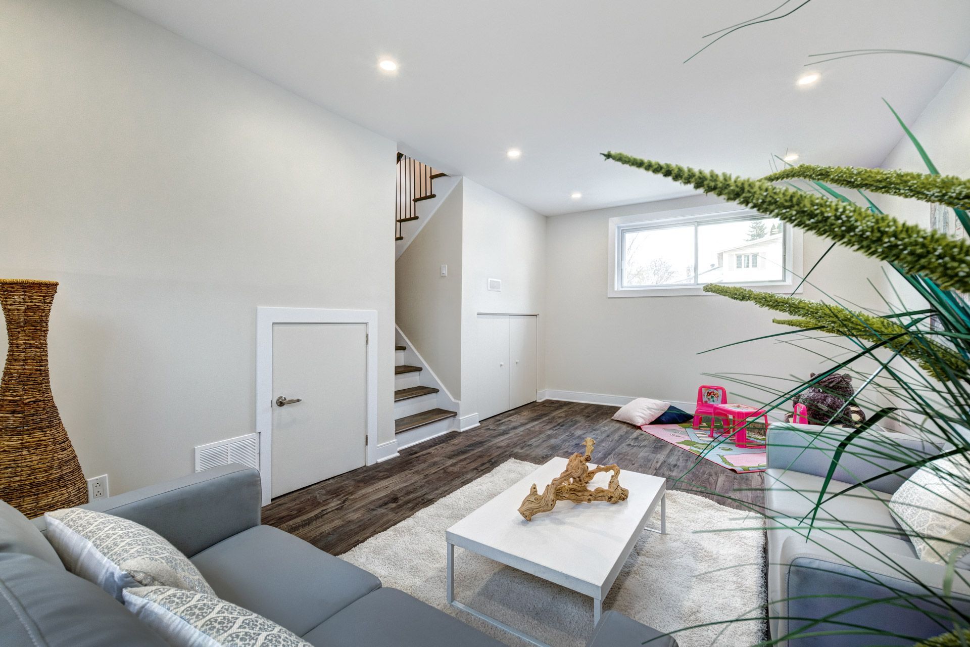 Bright living room with gray sofa, white coffee table, stairs, and large plant near a window