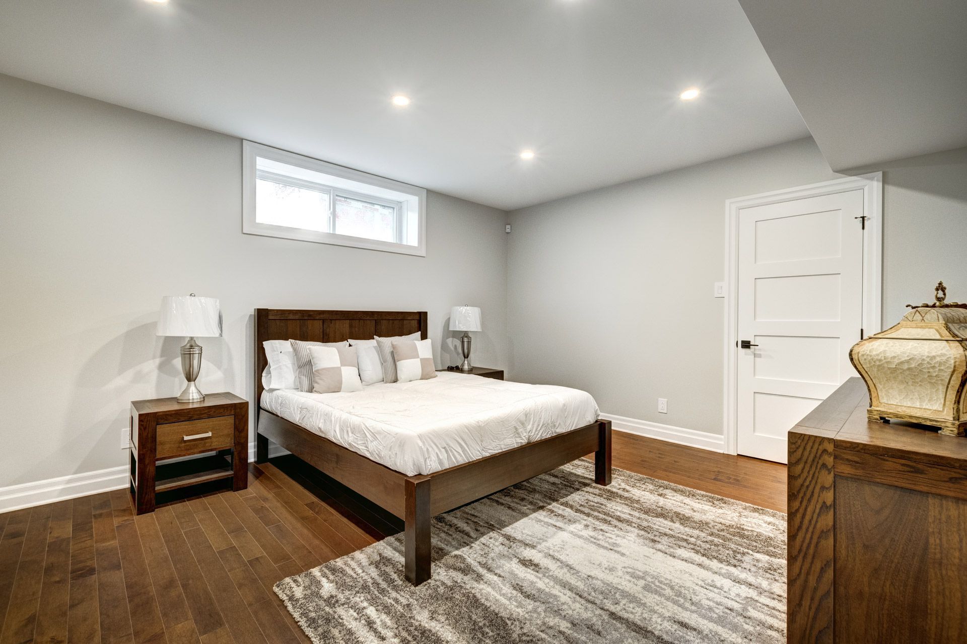 Bright bedroom with a wooden bed, white walls, lamps, rug, and dresser under recessed lights