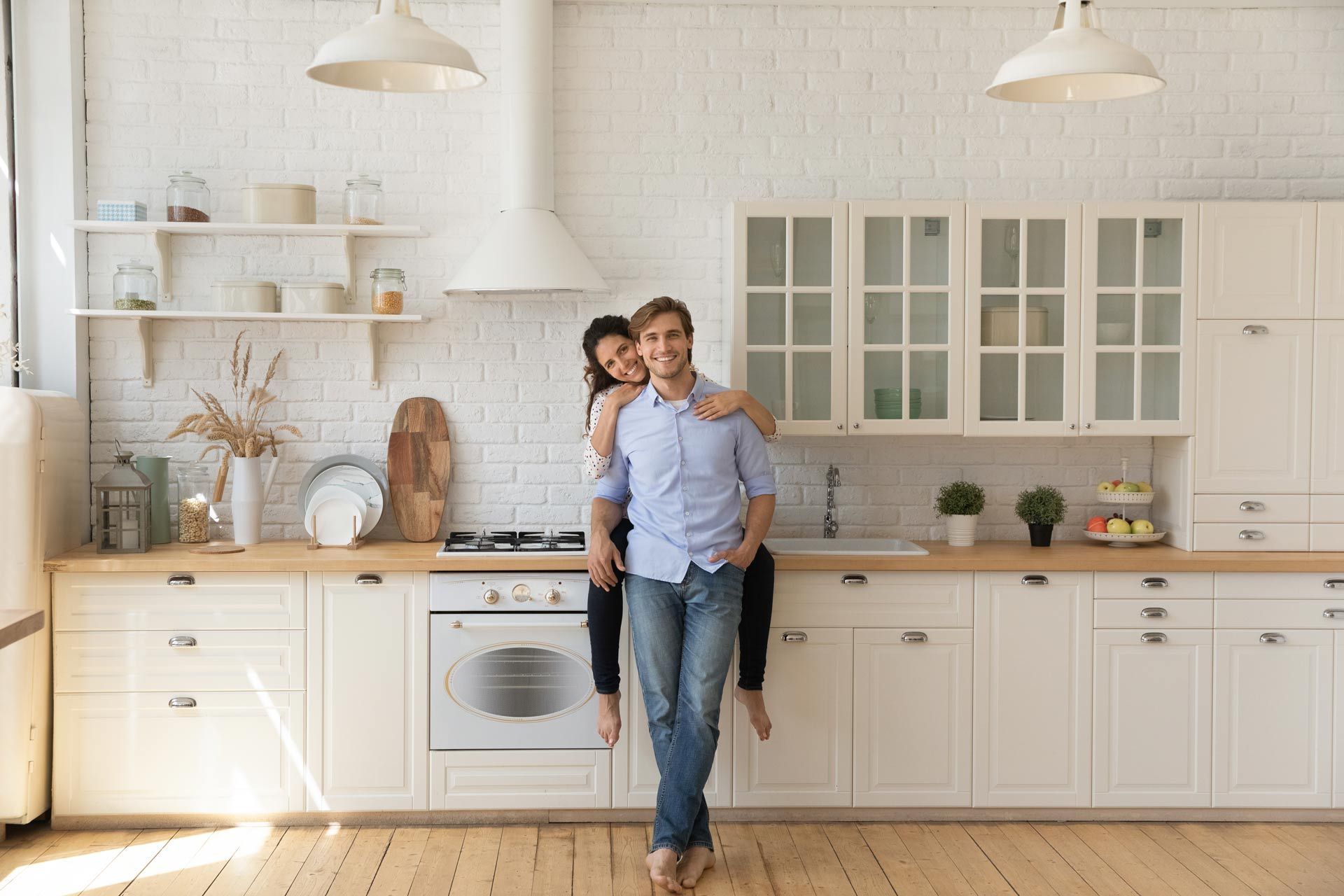 Couple standing in a bright white kitchen, one leaning on the other, with cabinets and hanging penda