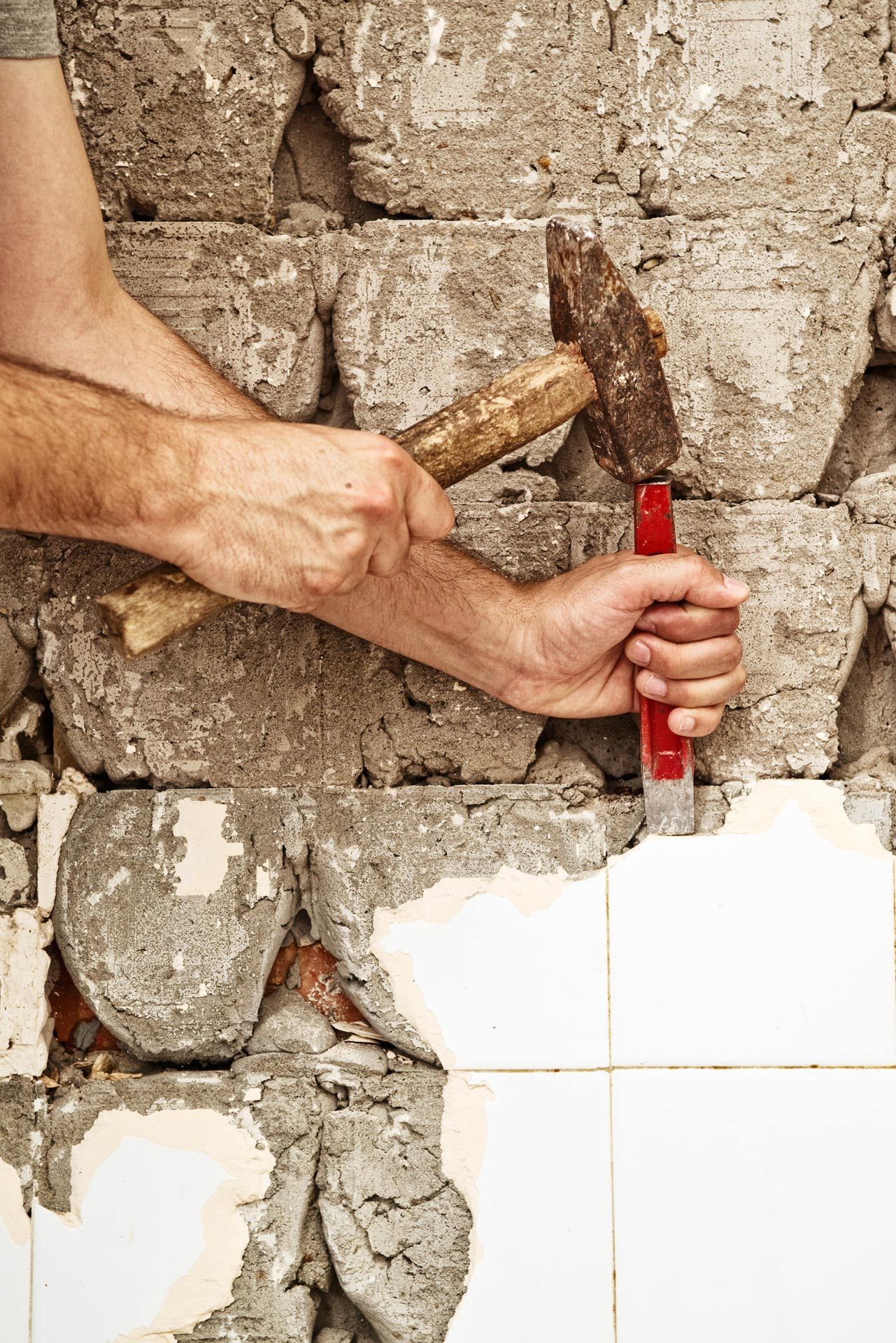 Hands with a hammer and chisel removing plaster from a brick wall beside white tiles. Hands with a hammer and chisel removing plaster from a brick wall beside white tiles.