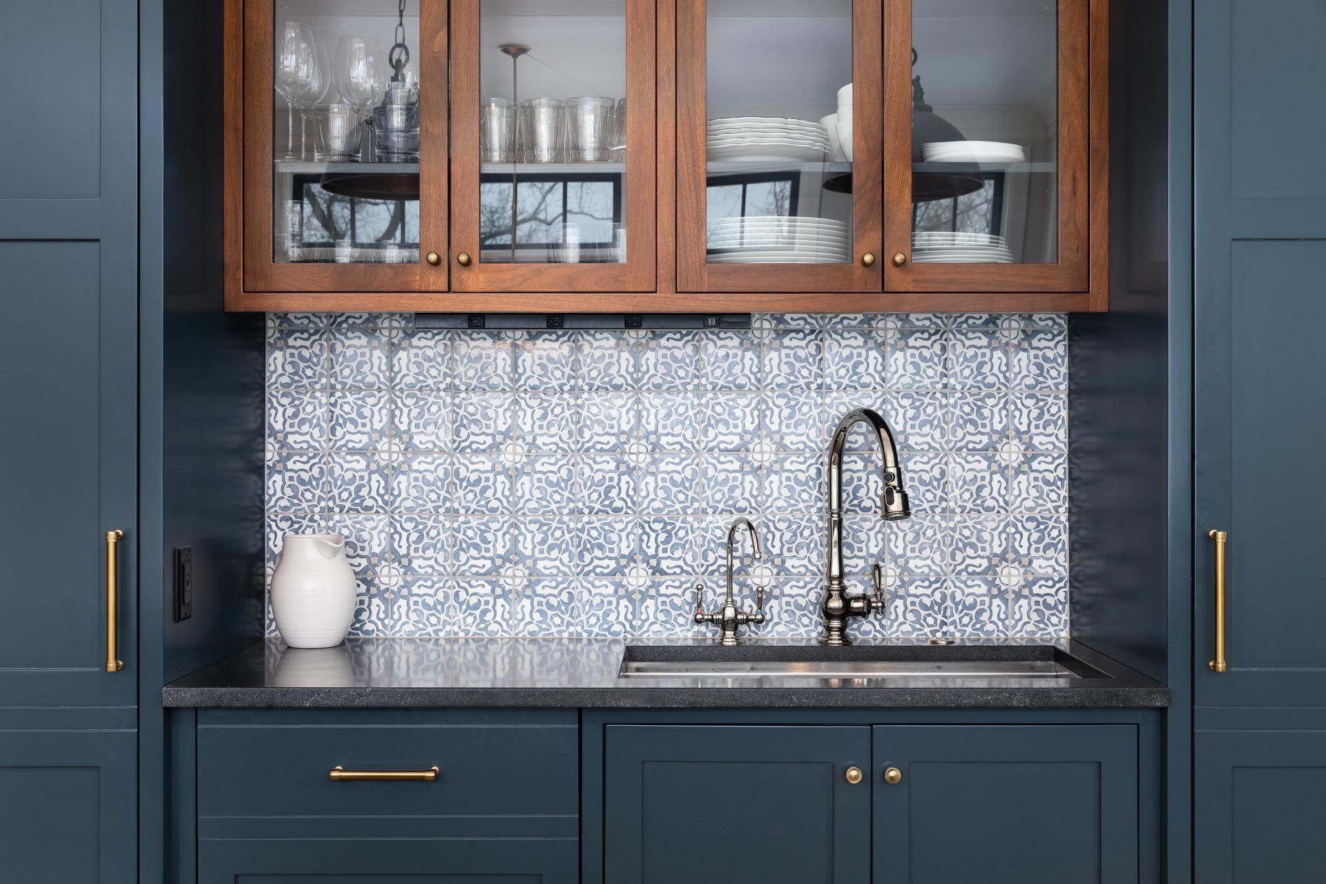 Navy kitchen cabinets with brass handles, wood-glass uppers, patterned tile backsplash, and sink faucet