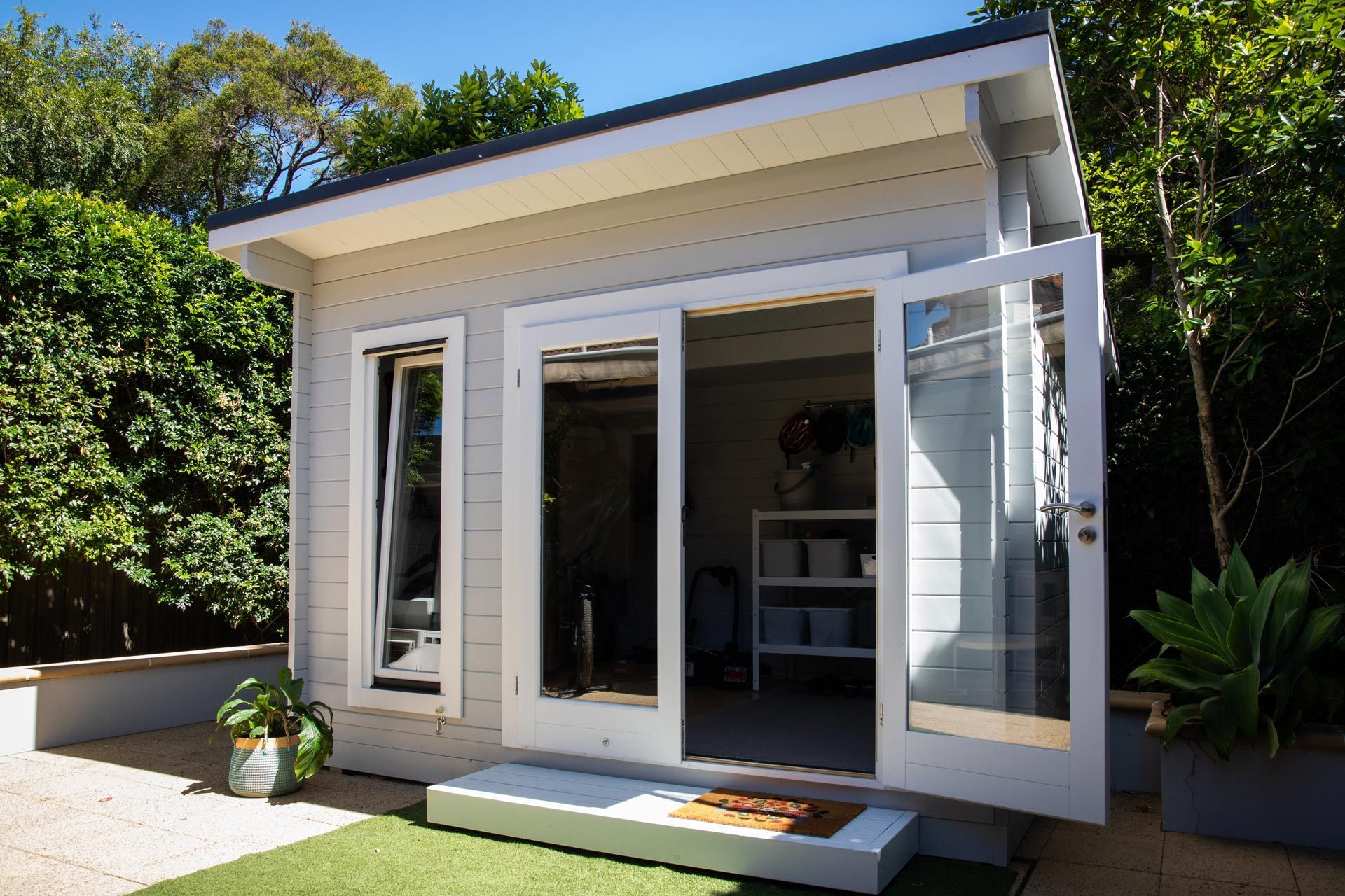 Small white garden shed with open doors and a front step, surrounded by greenery in bright sunlight
