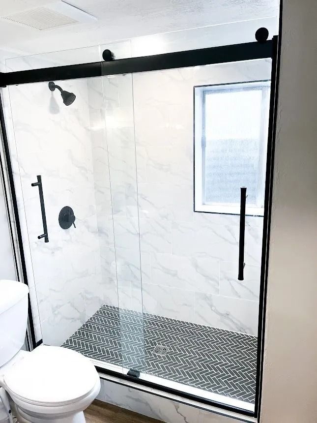 Modern glass walk-in shower with black hardware, white marble walls, and a pebble tile floor.
