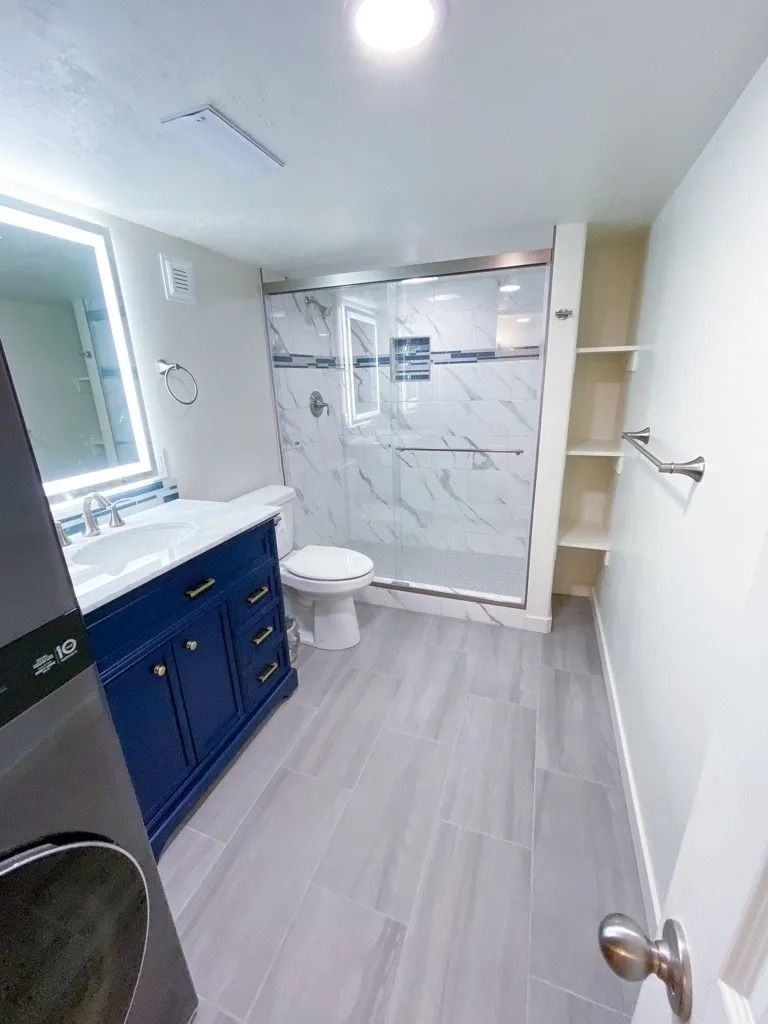 Modern bathroom with blue vanity, white marble shower, toilet, and gray wood-look floor.