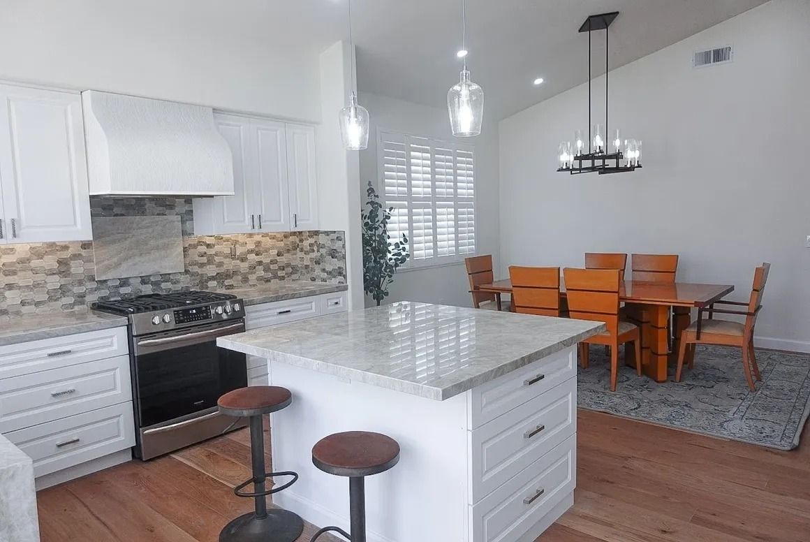 Bright kitchen with white cabinets, stone backsplash, island stools, and adjacent dining area with wooden table and chairs