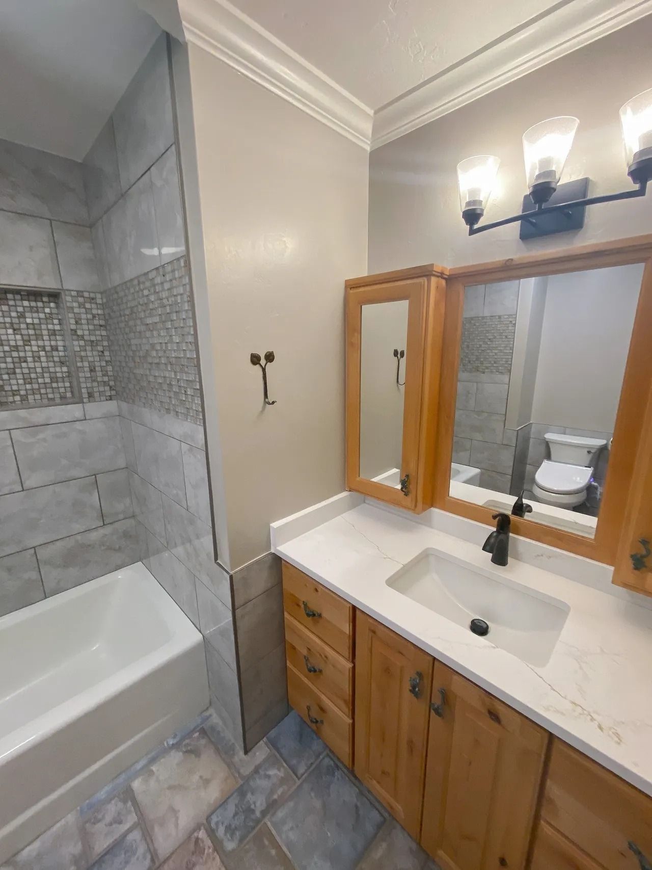 Bathroom vanity with mirror lights, wooden cabinets, white countertop, and adjacent tiled shower with tub.