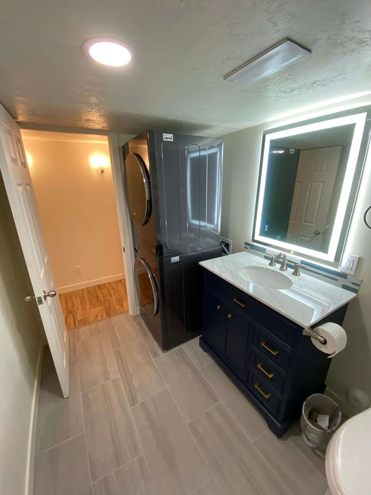 Modern bathroom with navy vanity, large lit mirror, glass shower door, and wood-look tile floor