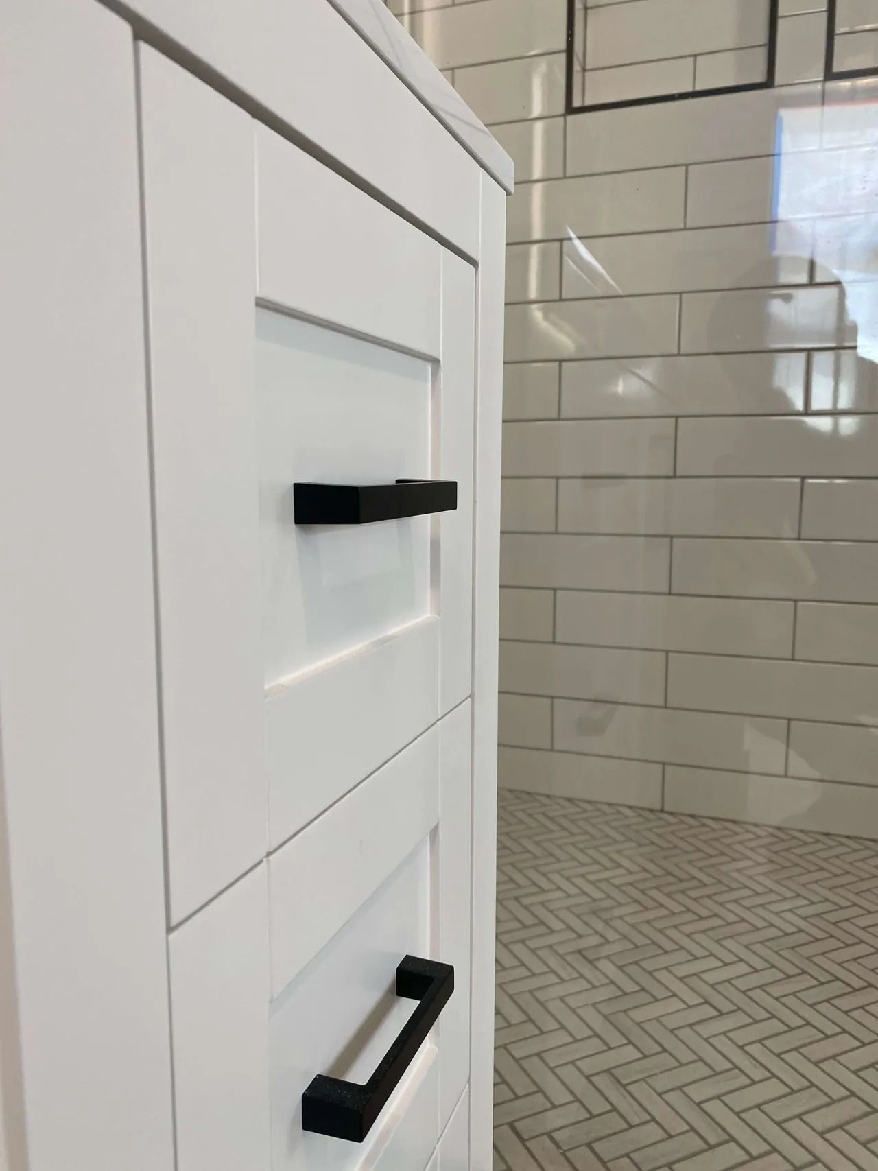 White bathroom vanity with black handles beside a tiled shower floor and white subway wall tiles