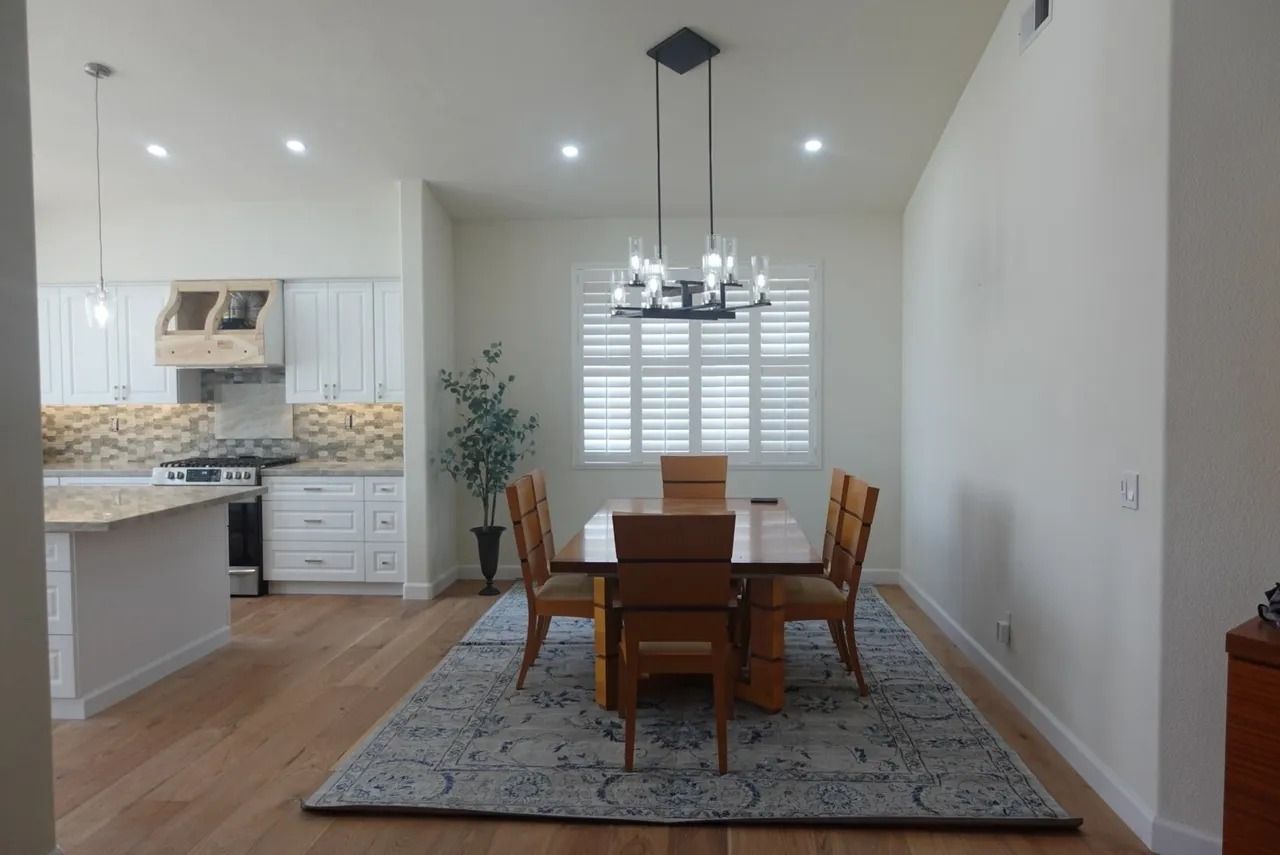Open dining area with wooden table, chandelier, gray rug, and bright white kitchen in the background
