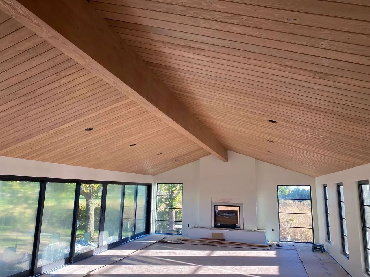 Modern living room with vaulted wood ceiling, large windows, and a central fireplace under construction