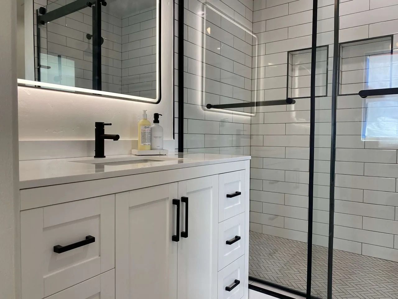 Modern bathroom vanity with white cabinets, black fixtures, and a glass-enclosed shower with white tile.